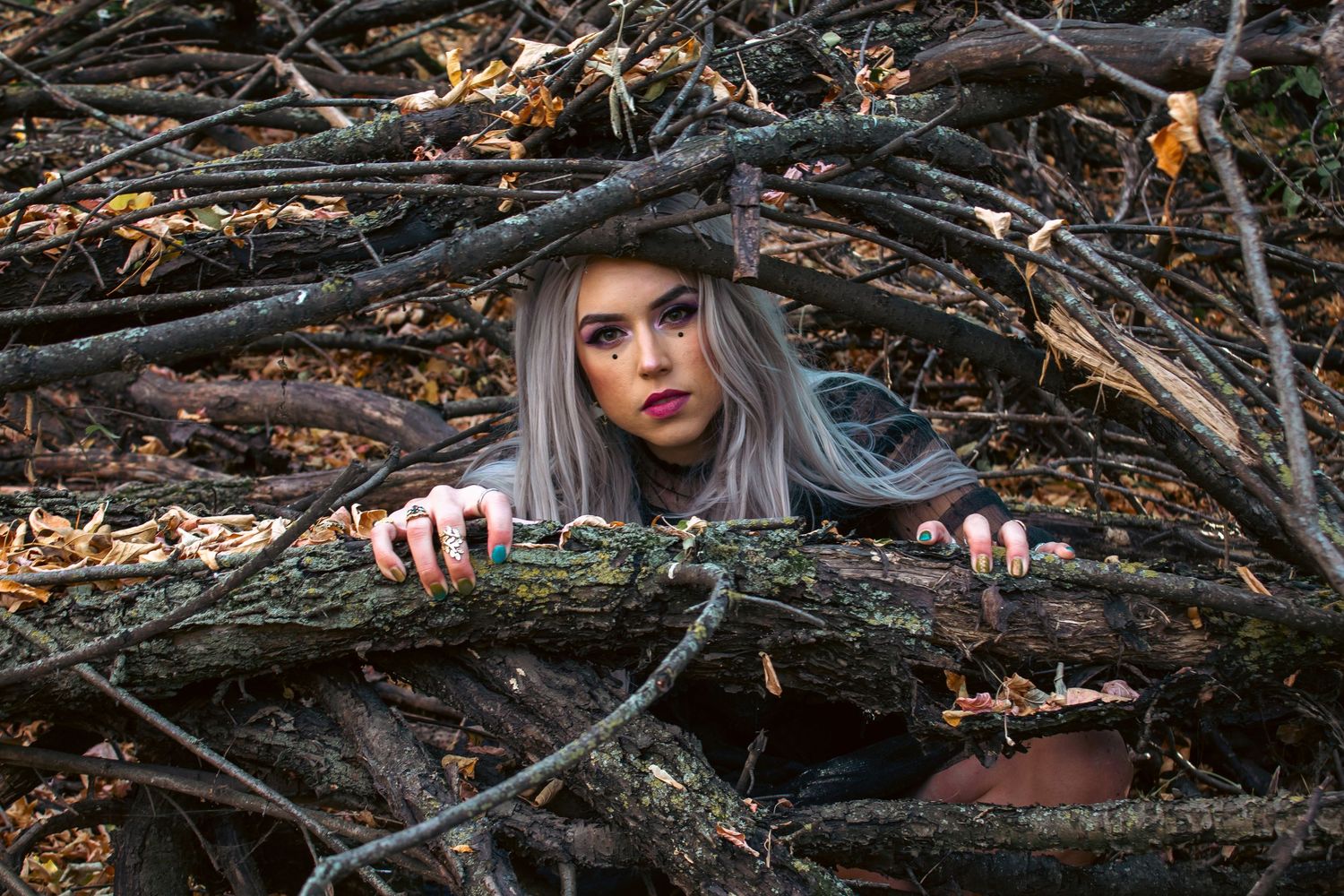 Woman with long silver hair and dark lipstick peers over a fallen mossy log, hands resting on the bark in a tangled autumn forest.