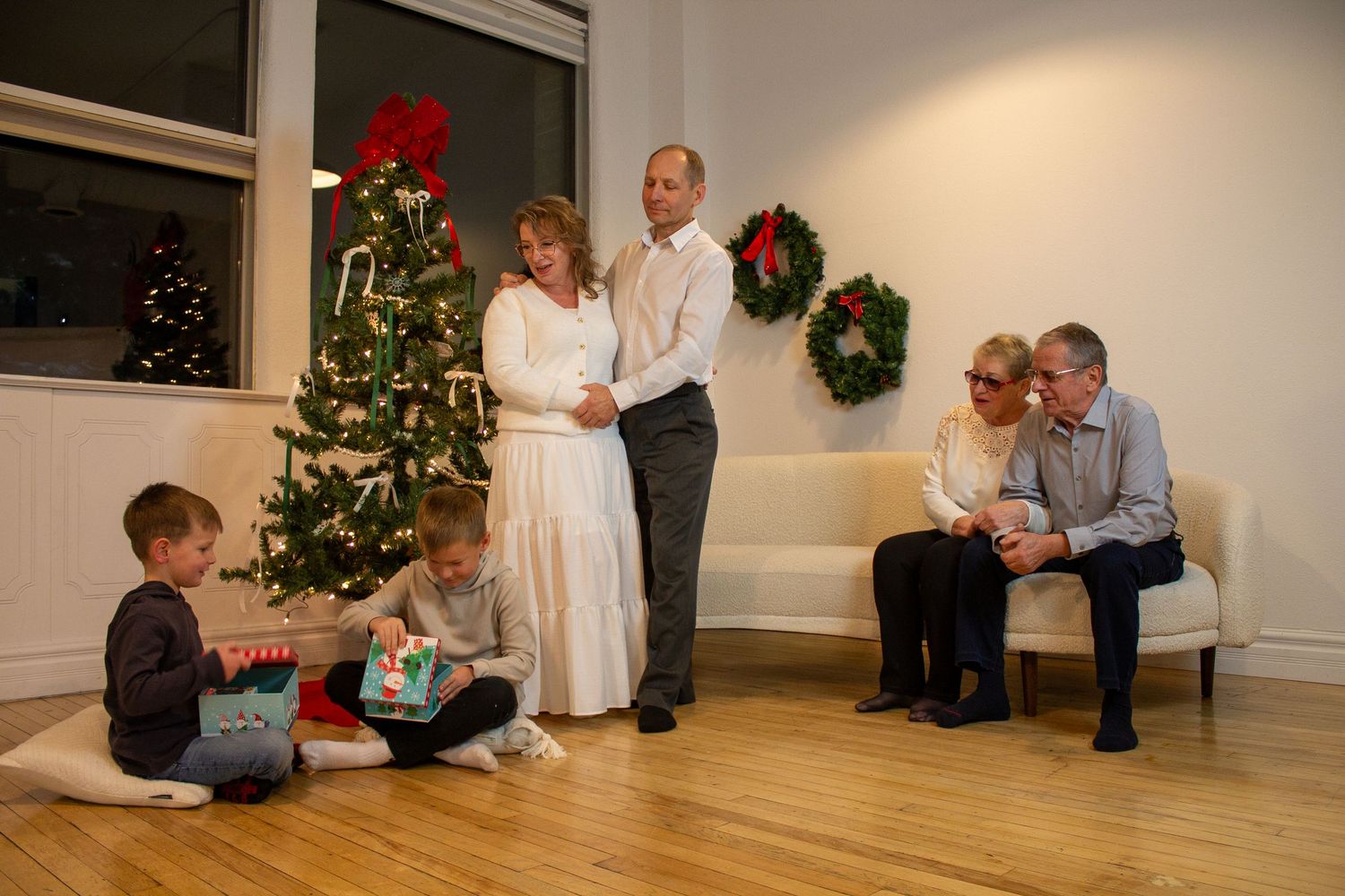 Multigenerational family gathers near a decorated Christmas tree and wreaths as two boys open gifts while parents and grandparents watch.