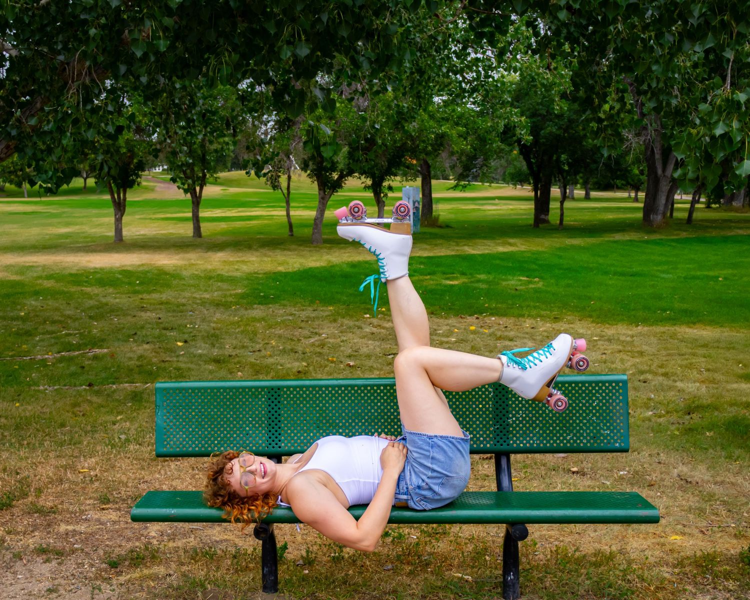 Woman in roller skates lies back on a green park bench with her legs raised, surrounded by grass and trees on a sunny day.