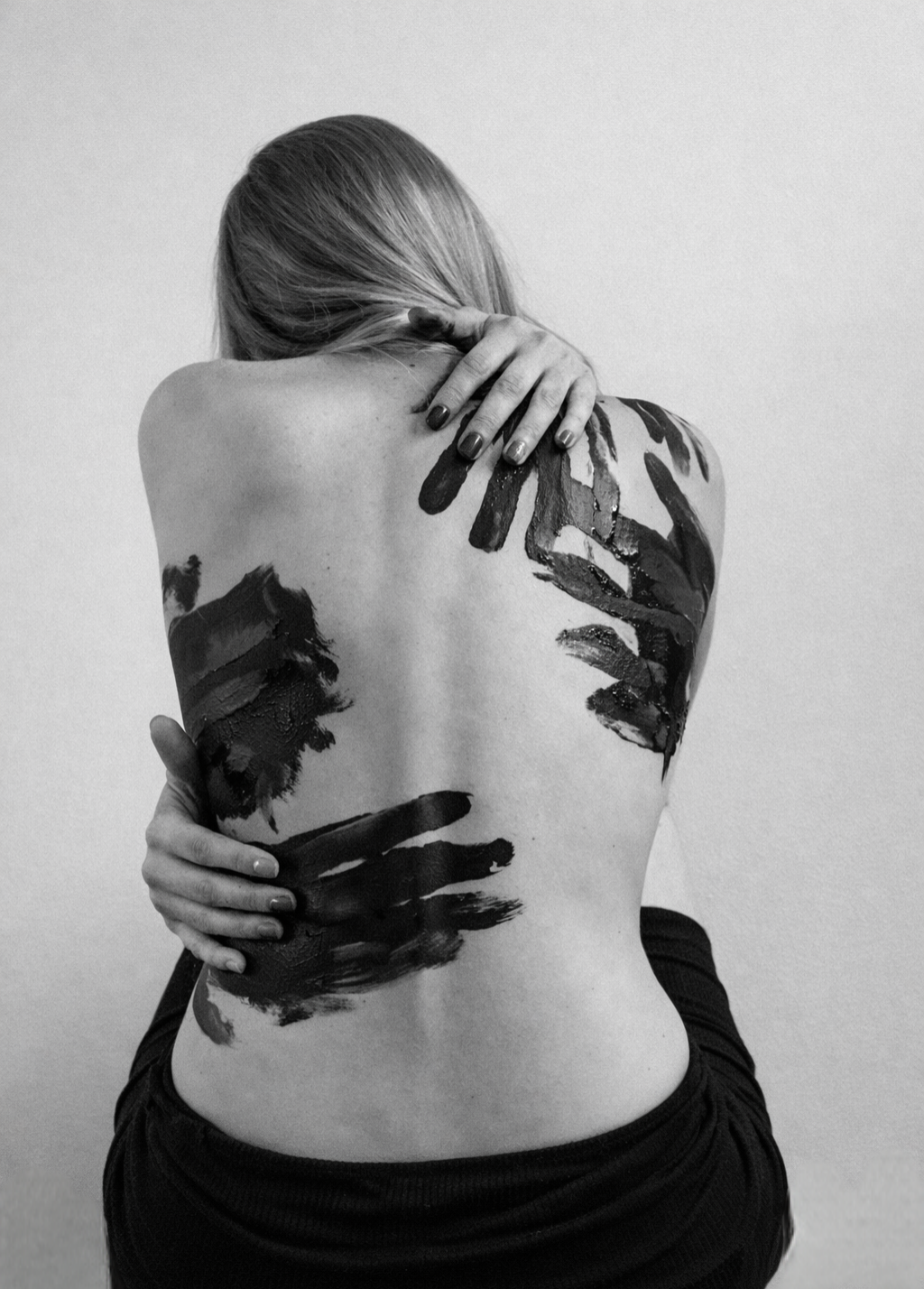 Black and white photo of a woman from behind, hugging herself, with dark painted handprints smeared across her bare back.