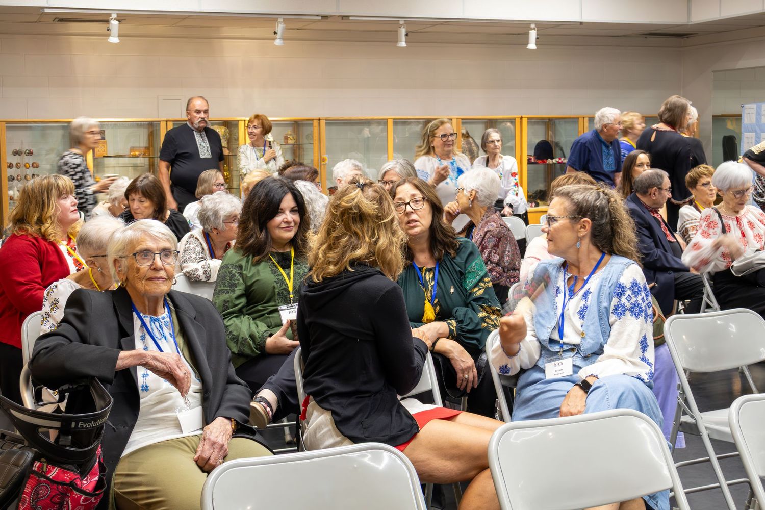 A diverse group of older adults sits and talks in rows of folding chairs at a community gathering in a bright indoor event space.
