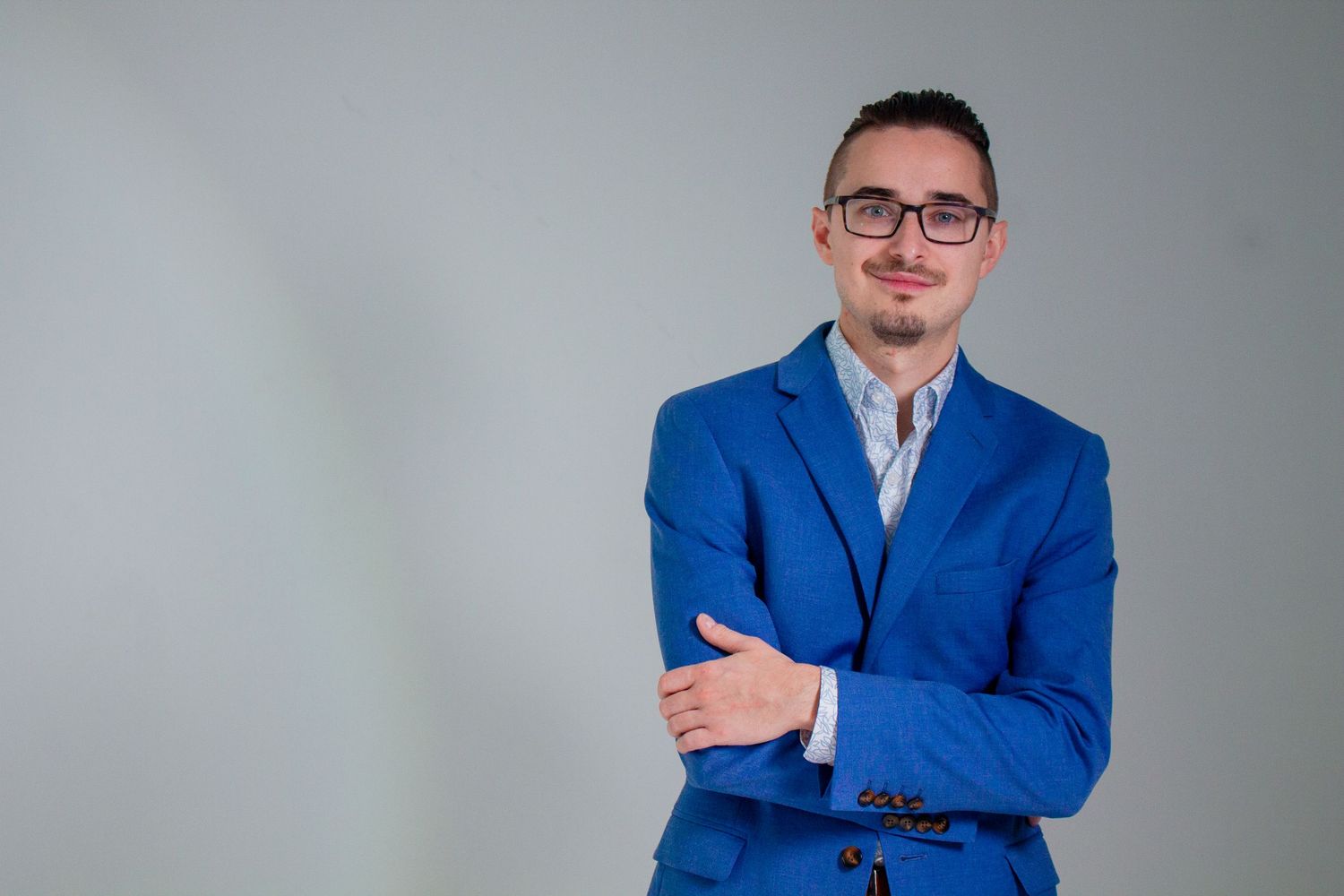 Smiling man with glasses and a goatee wears a bright blue blazer and light shirt, posing with arms crossed against a gray studio background.