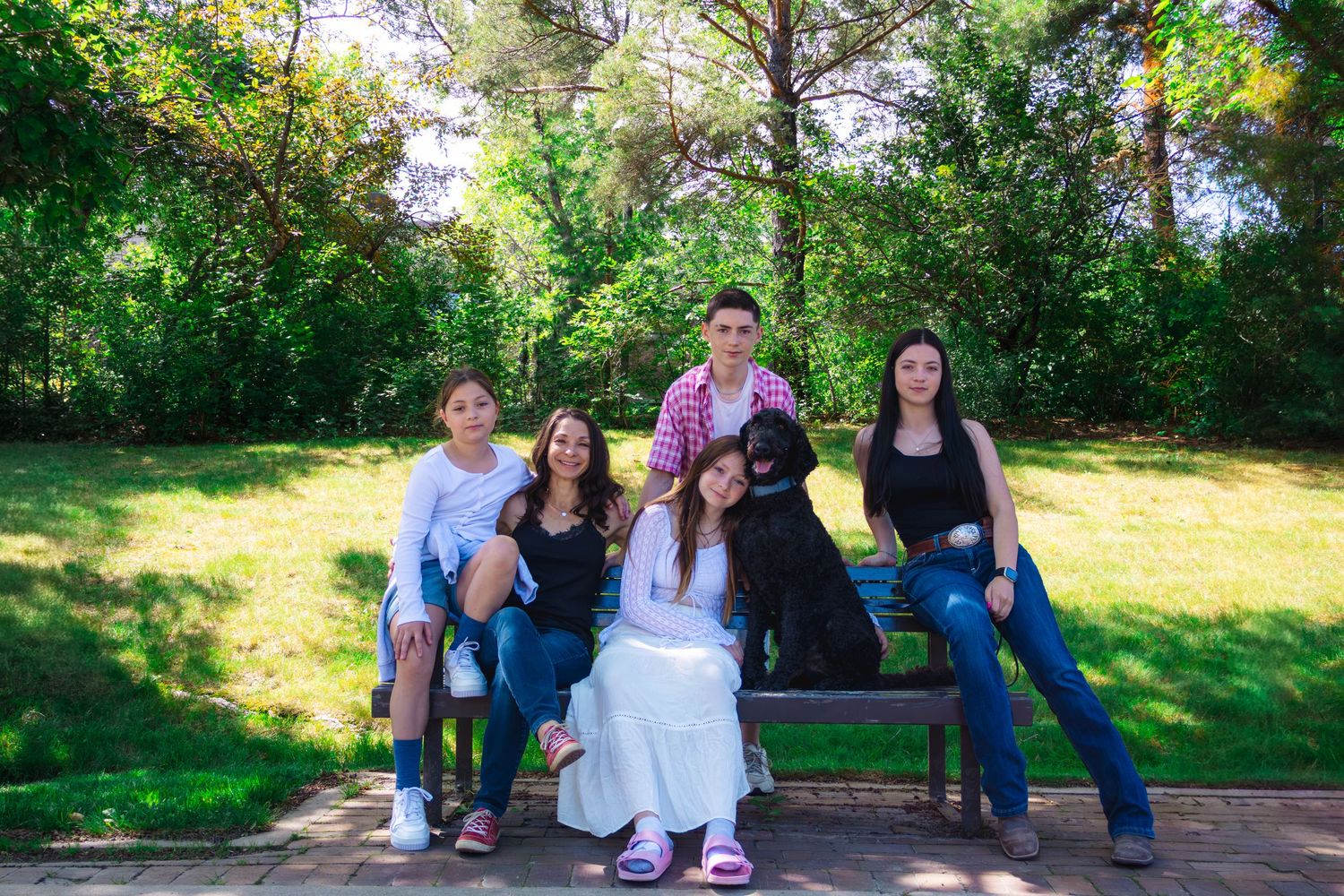 Five young people and a black dog pose together on a wooden park bench surrounded by green trees and grass on a sunny day.