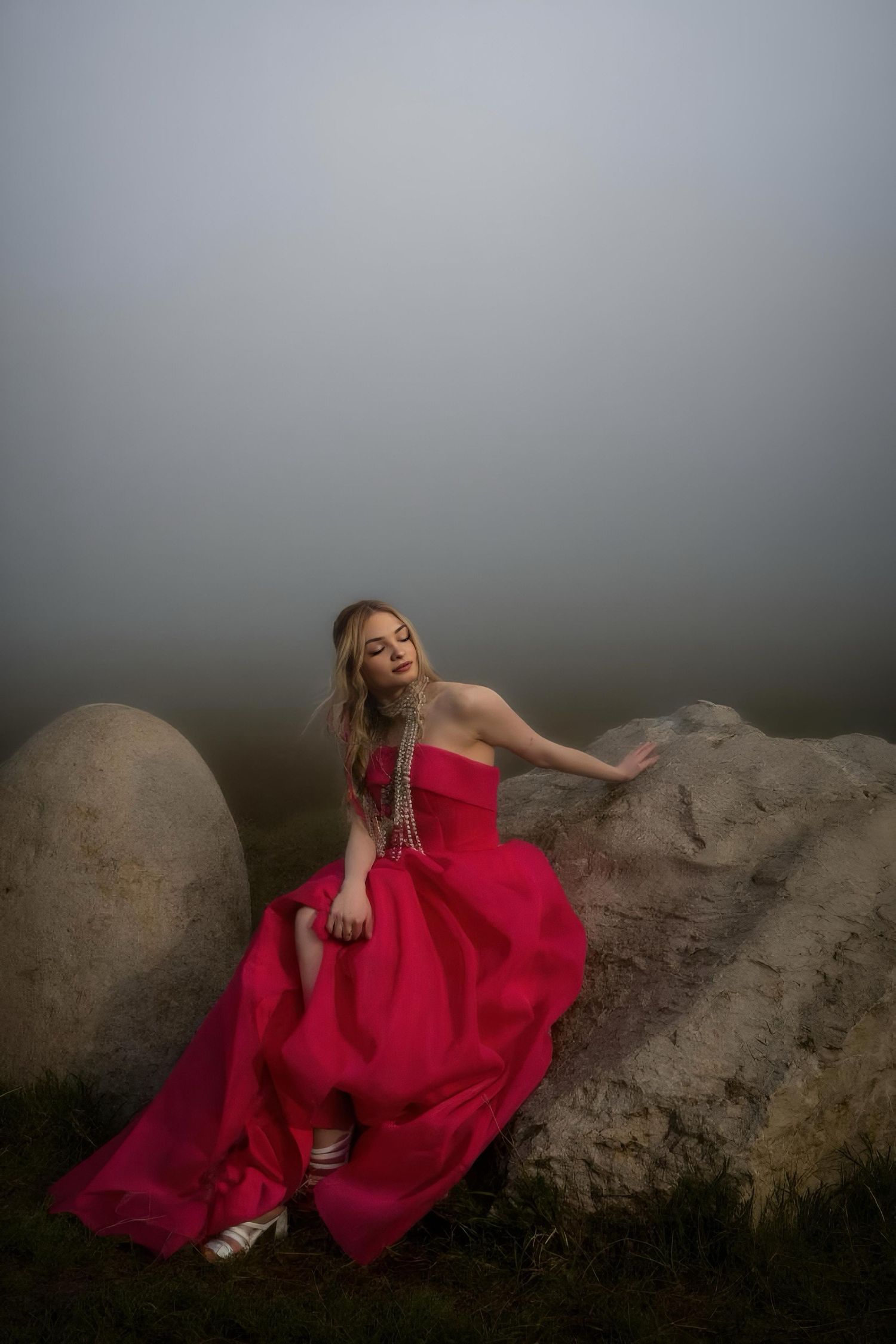 Woman in a flowing magenta strapless gown sits on a large boulder in a foggy, moody landscape with her eyes closed.