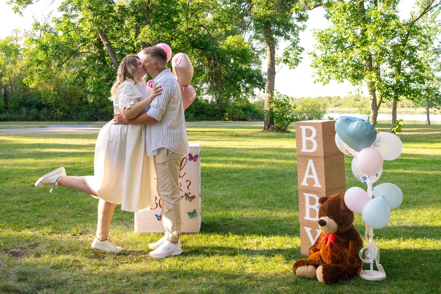 Couple embracing and kissing in a sunny park beside BABY letter blocks, pastel balloons and a teddy bear at a gender reveal photo shoot.