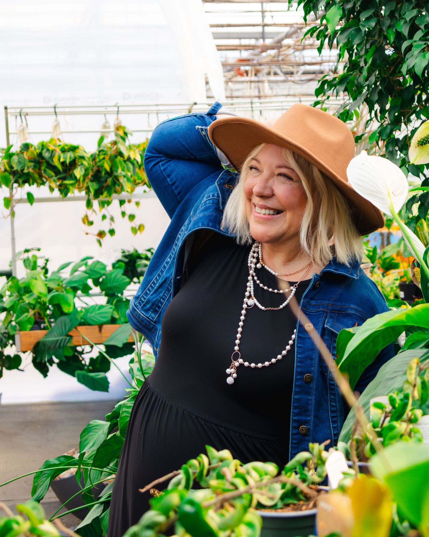 Smiling woman in a tan wide-brim hat, denim jacket and long beaded necklace stands among lush green plants in a bright greenhouse.