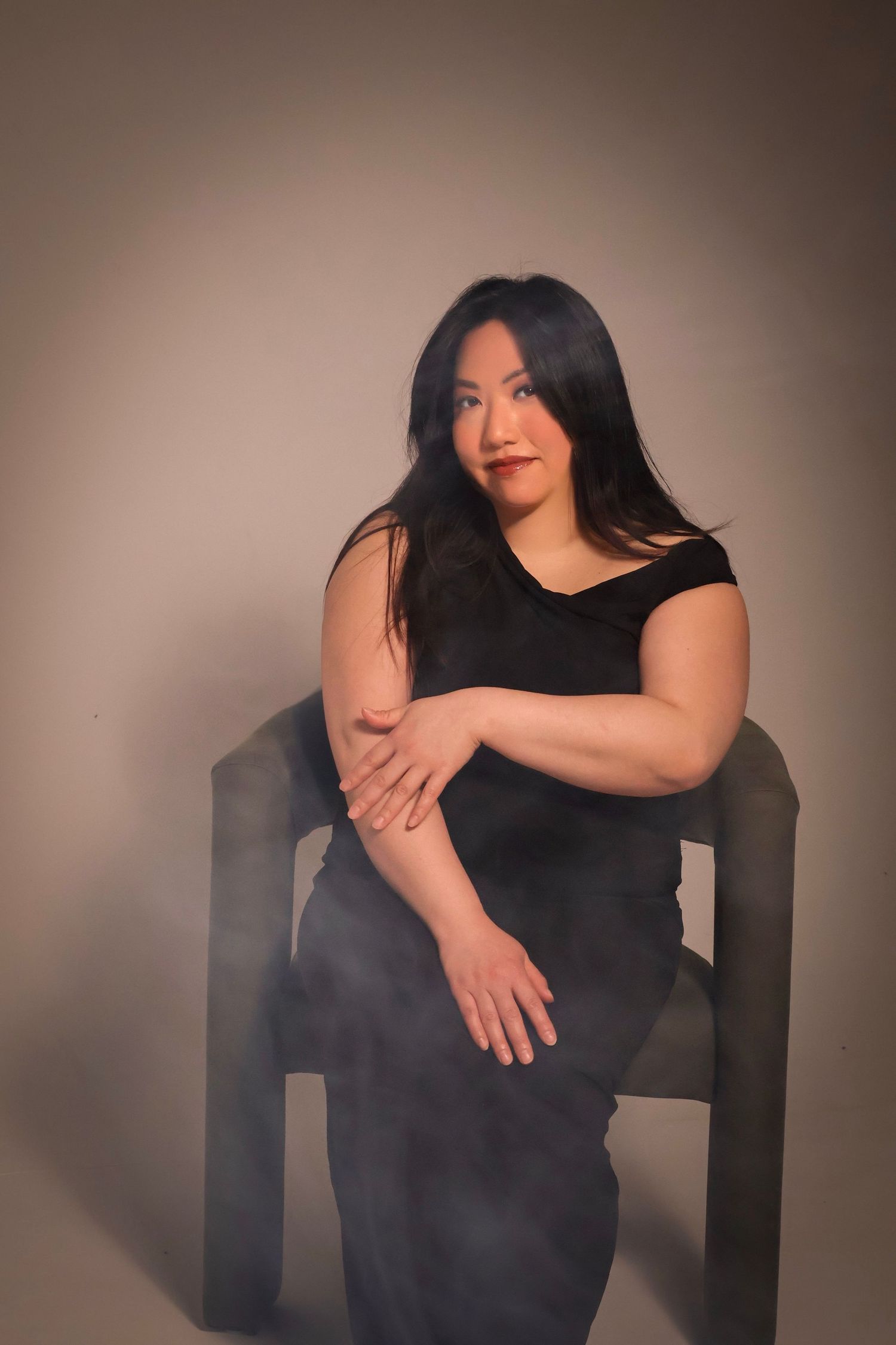 Woman with long dark hair in a black off-the-shoulder dress sits on a gray modern chair, arms crossed, in a soft-lit studio portrait.