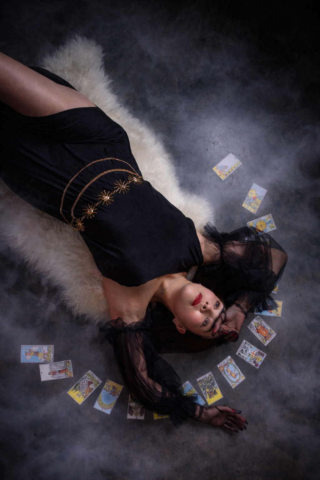 Woman in a black dress lying on a white fur rug surrounded by scattered tarot cards in a moody, smoky studio setting.