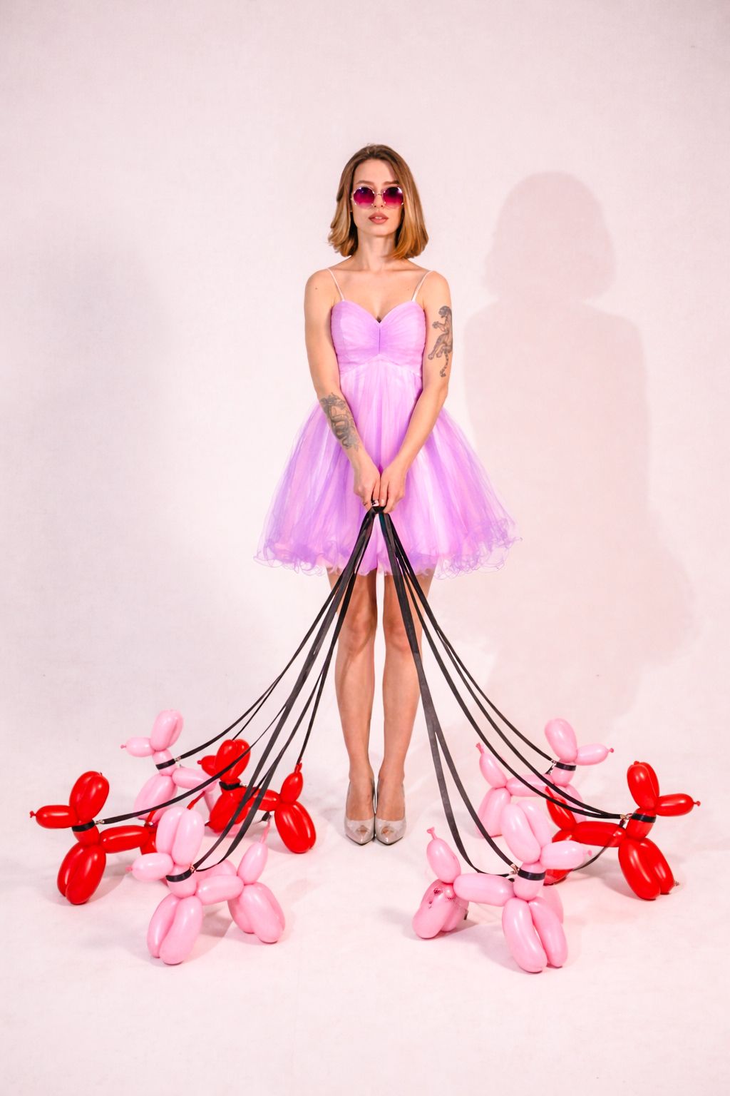 Woman in a pink tulle dress and red sunglasses holds leashes attached to pink and red balloon dogs against a white studio backdrop.