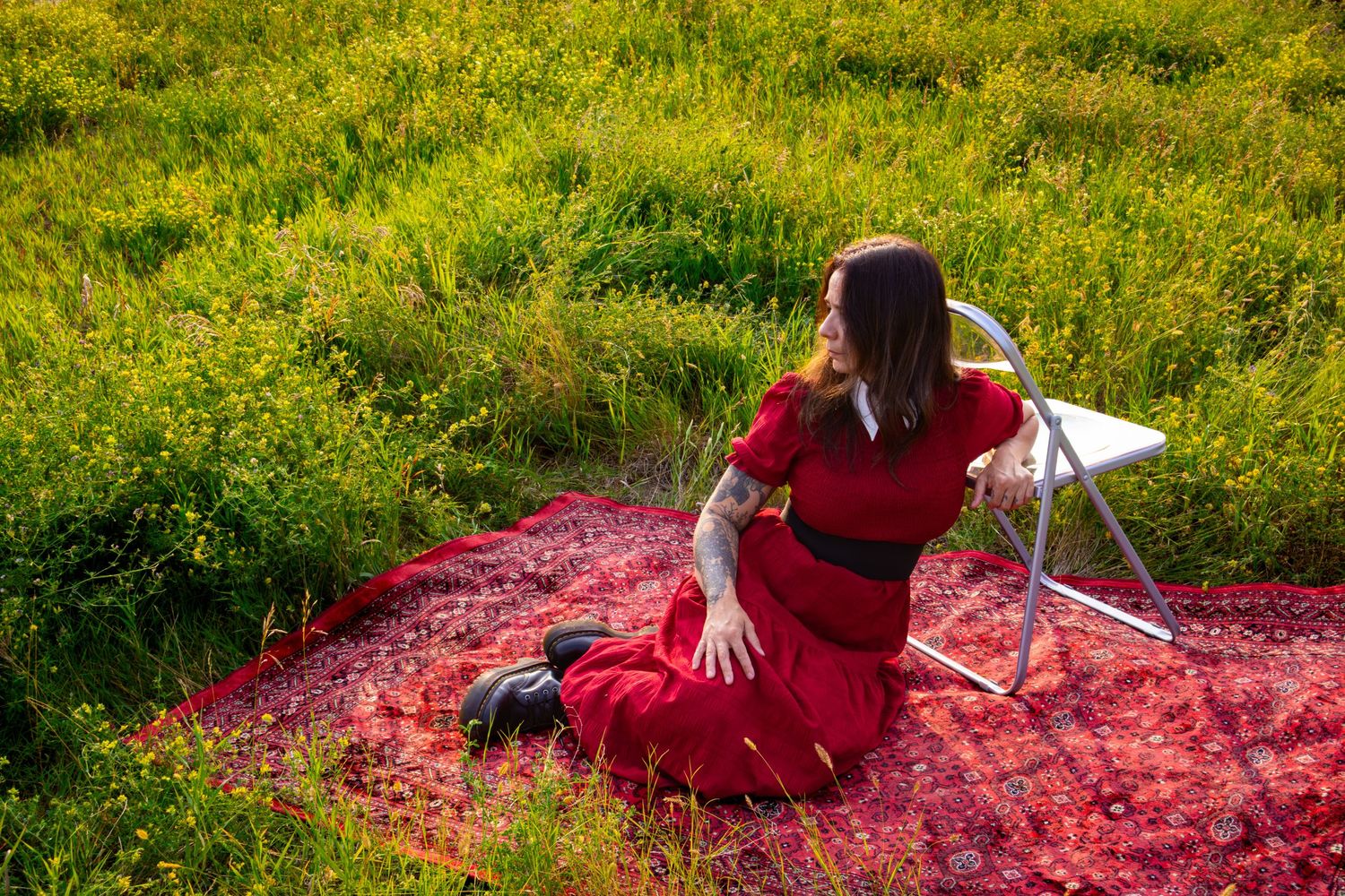 Woman in a red dress and black boots sitting on a red patterned blanket in a sunlit green meadow beside a white folding chair.