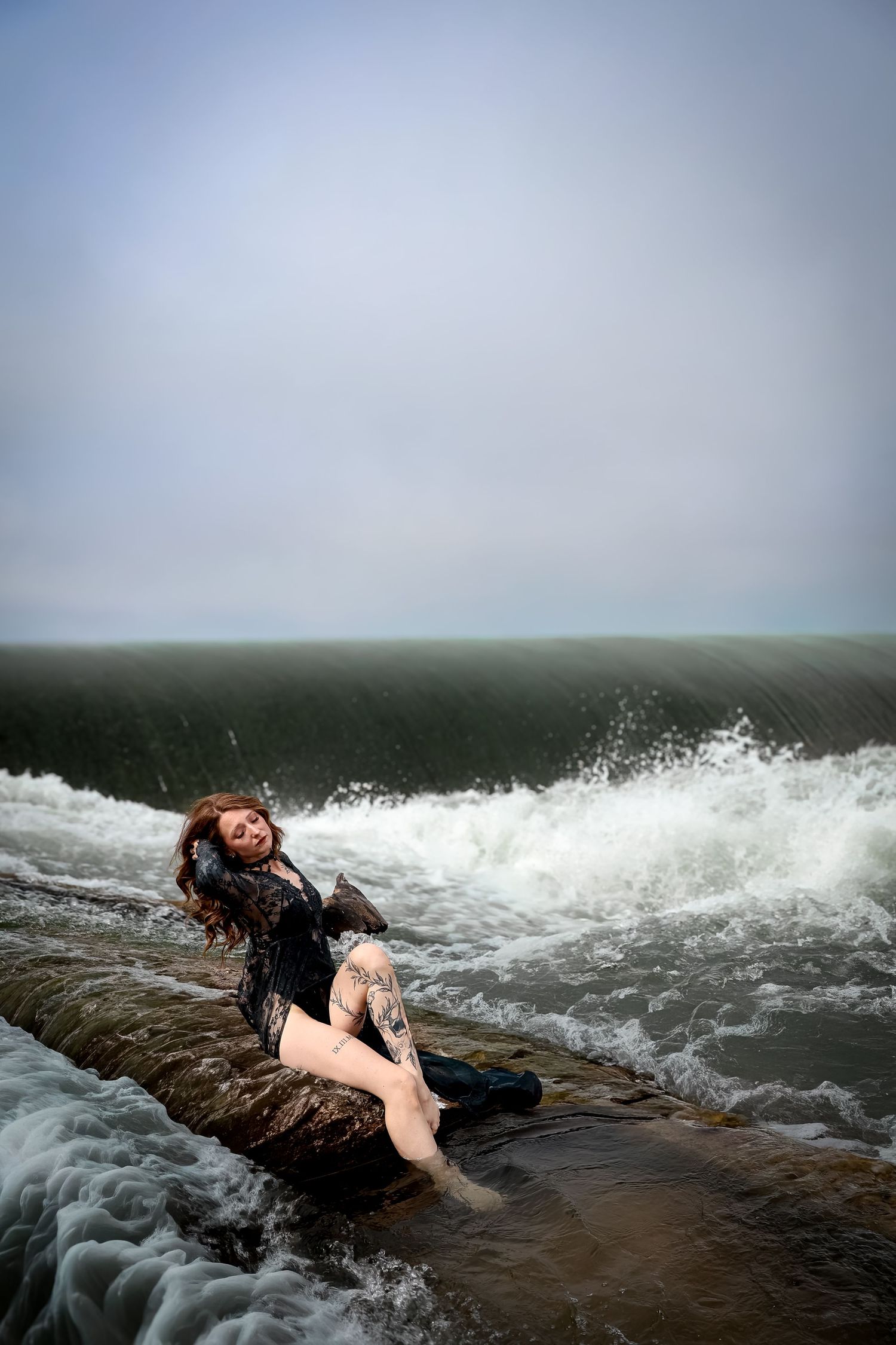 Woman with auburn hair and leg tattoos sits on a mossy ledge as ocean waves crash around her under a soft gray sky.