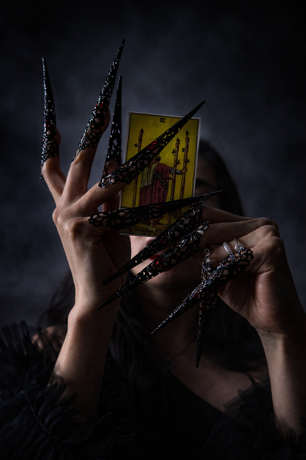 Woman in dark clothing holds a yellow tarot card between long ornate black claw-style fingertip nails against a moody smoky background.