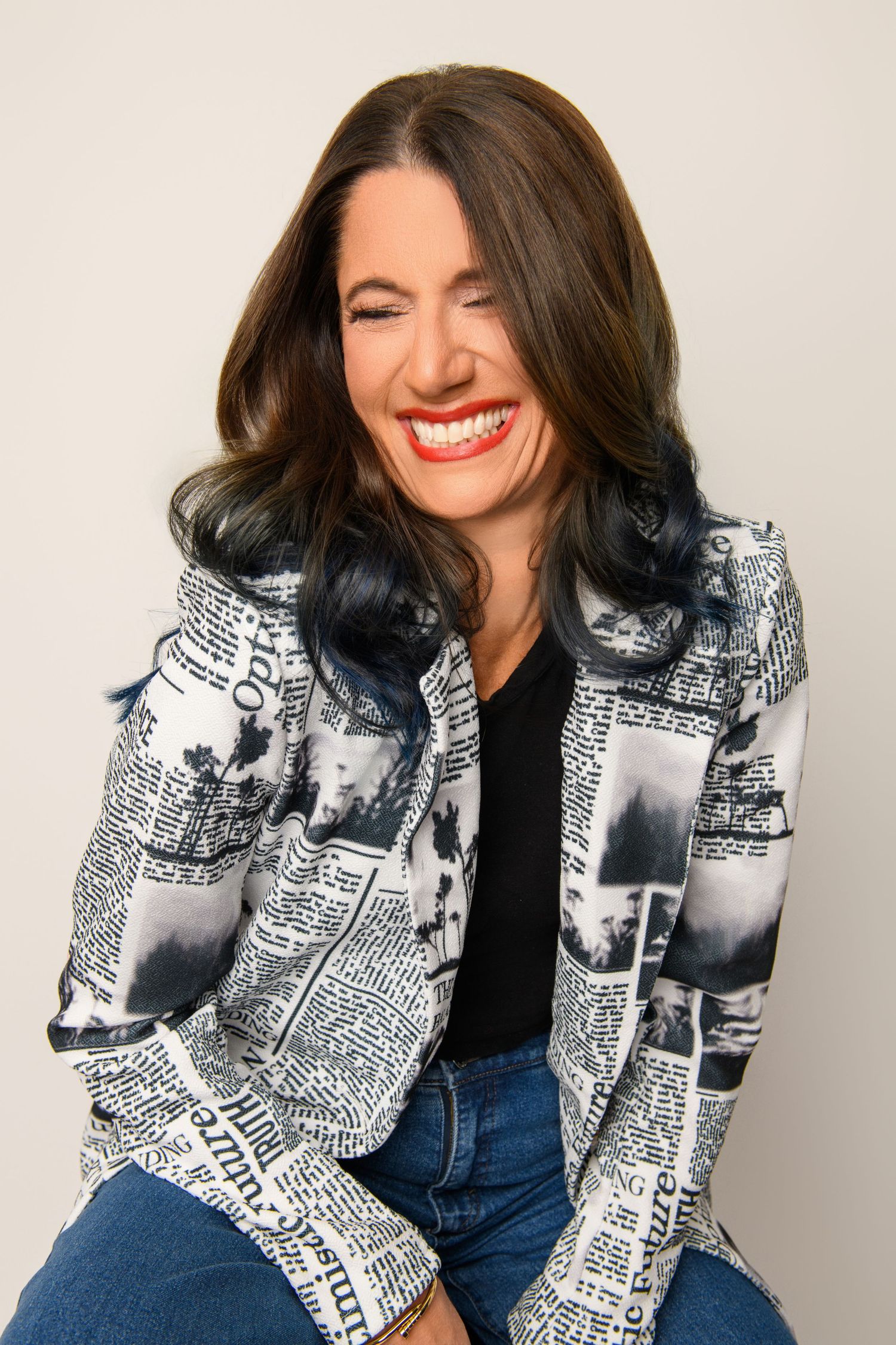 A person wearing a black and white newspaper print jacket and blue jeans laughs joyfully while sitting.