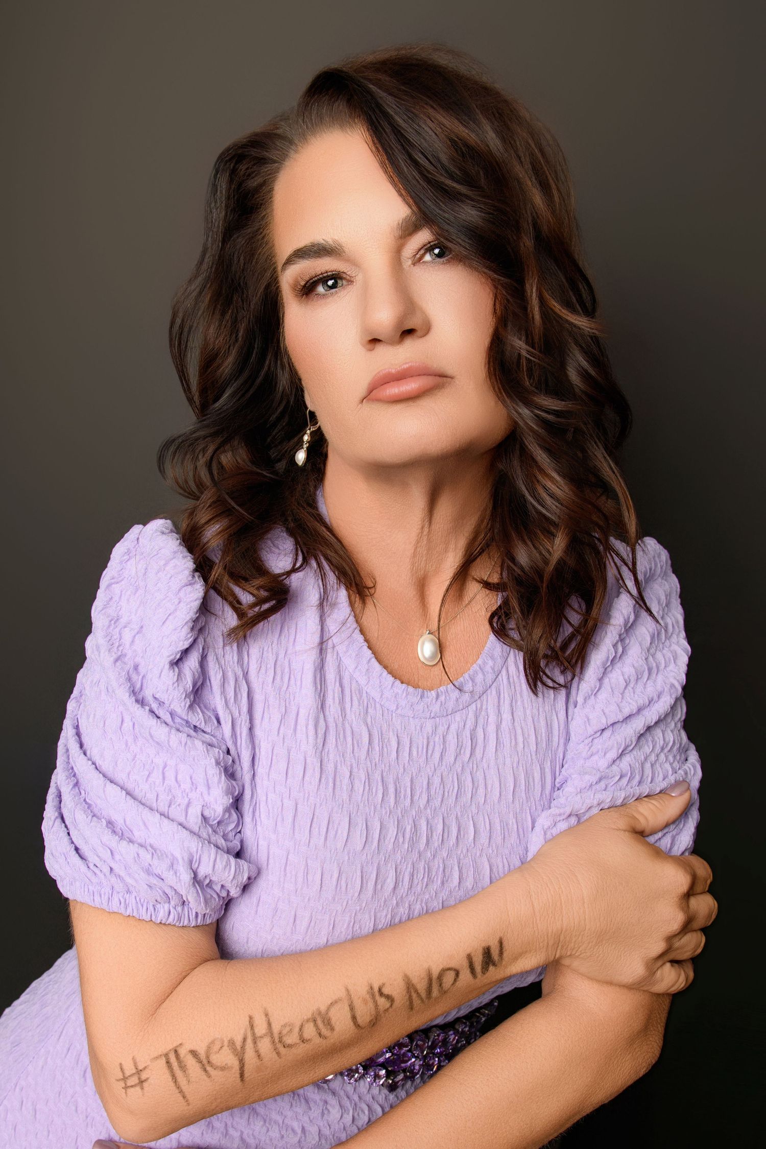 Portrait of woman in lavender top with wavy hair and delicate jewelry against dark background.