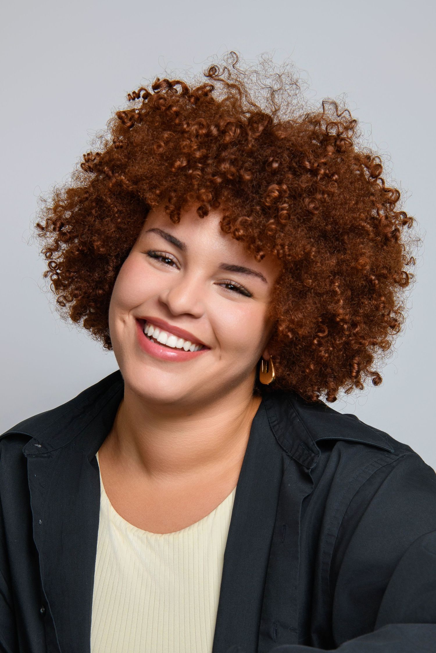 Professional headshot portrait of someone with voluminous natural curly brown hair wearing a black blazer and light top.