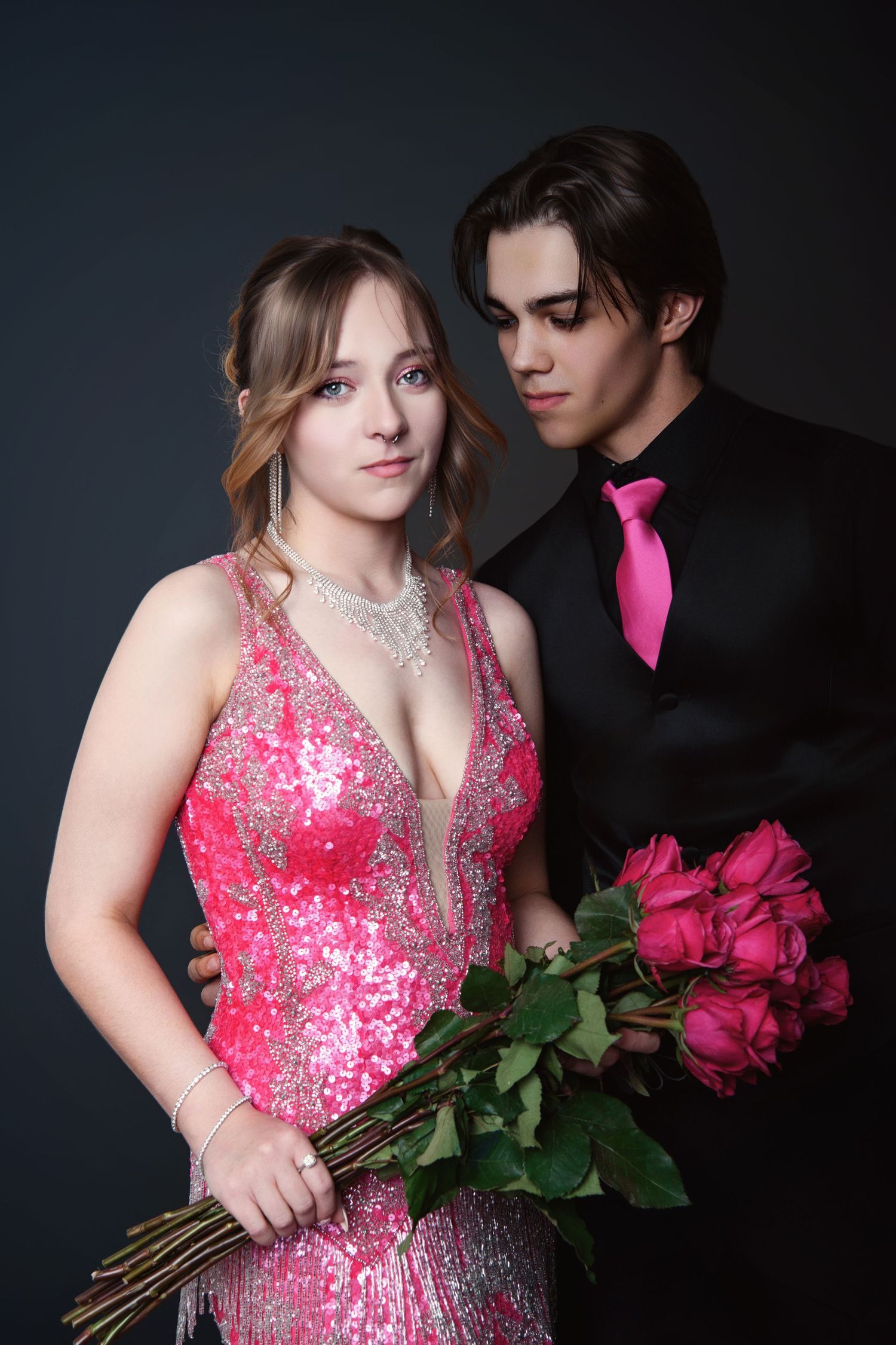 Elegant couple poses for prom photo with red roses, woman in pink sequin gown and man in black suit.