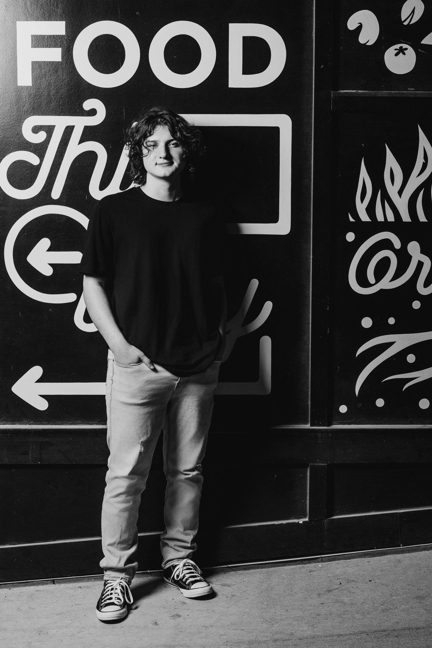Casual portrait of person in black shirt and jeans standing against wall with food signage.