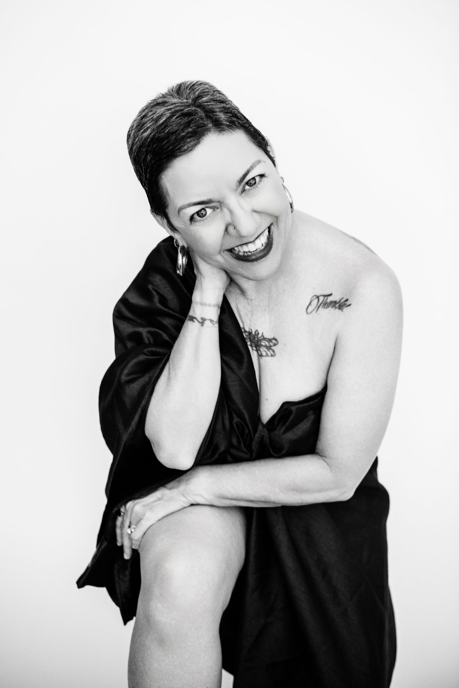 Artistic black and white photo of a person in black dress smiling confidently with visible tattoos.