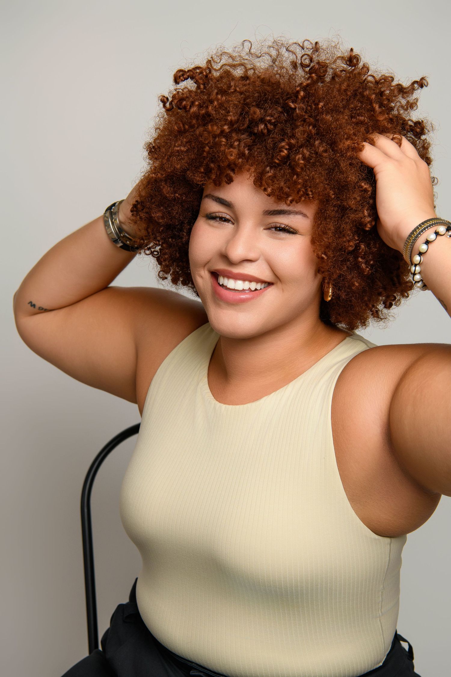 Happy person with curly brown hair wearing a cream sleeveless top poses with hands running through their hair.
