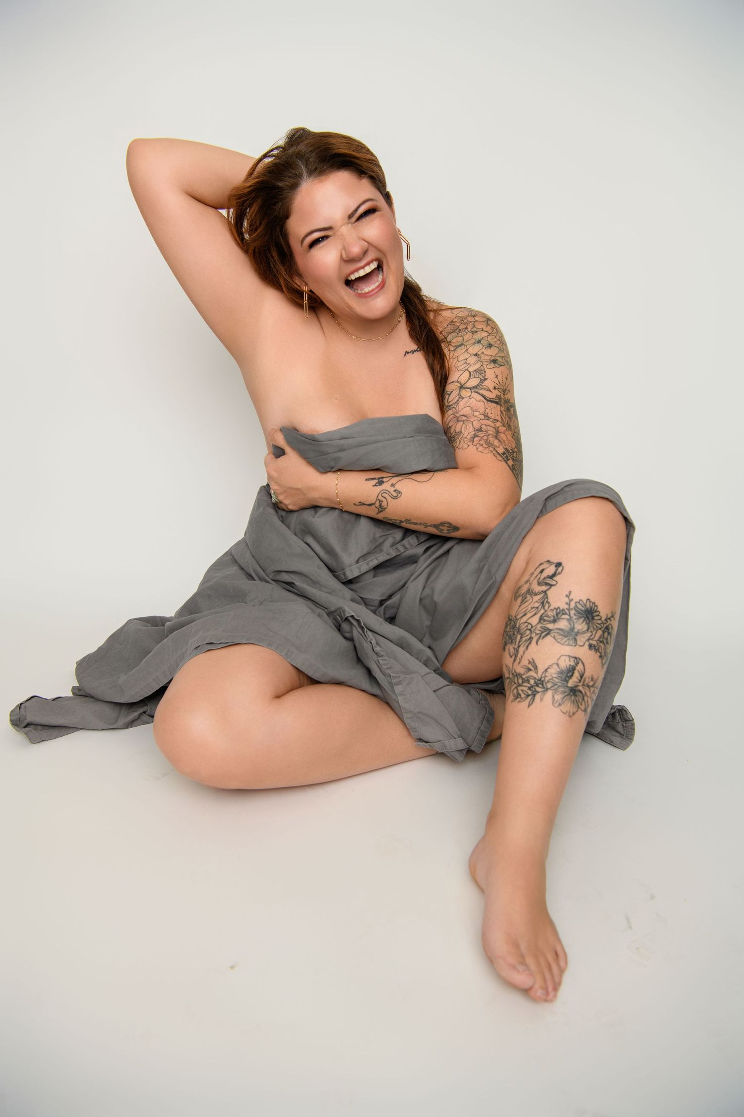 Person wearing gray dress sitting on white background showing tattoos while laughing joyfully.