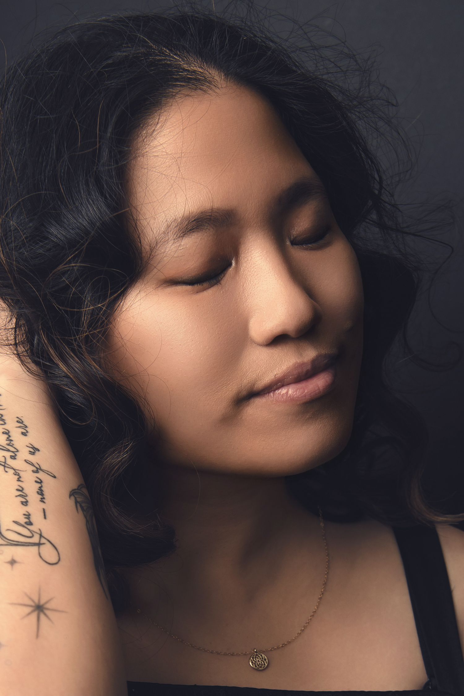 Artistic portrait of someone in soft lighting with closed eyes, dark hair, and a delicate necklace against dark background.