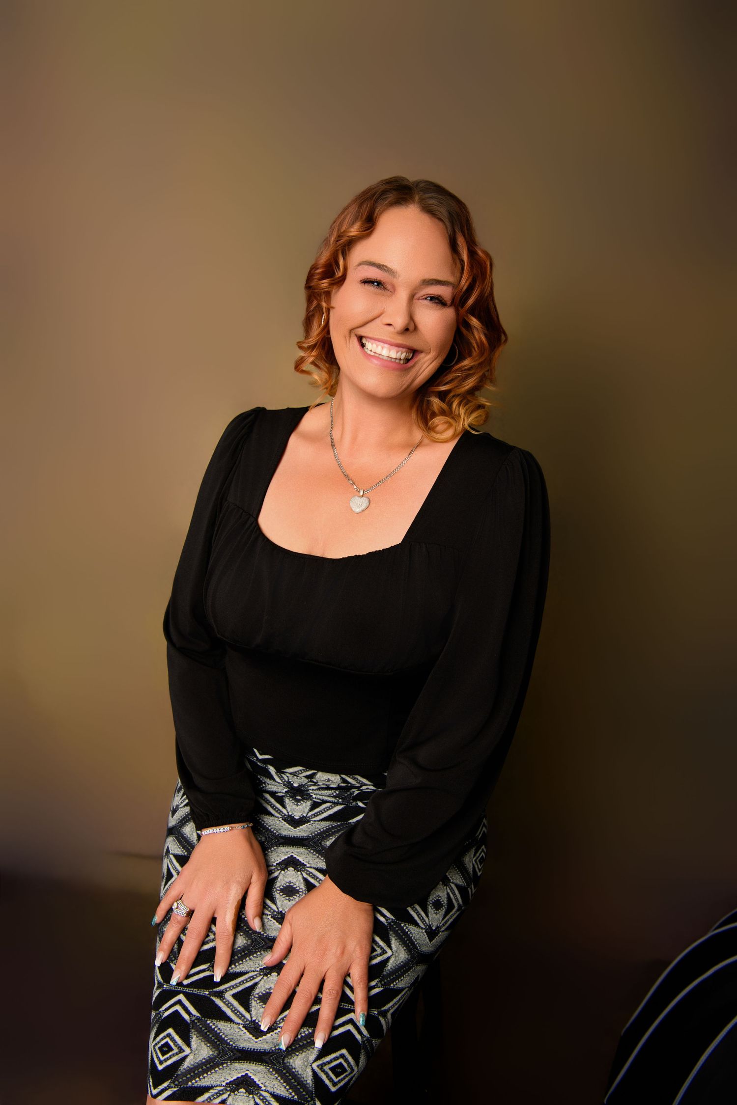 Professional portrait of someone wearing a black top and patterned skirt against warm background.