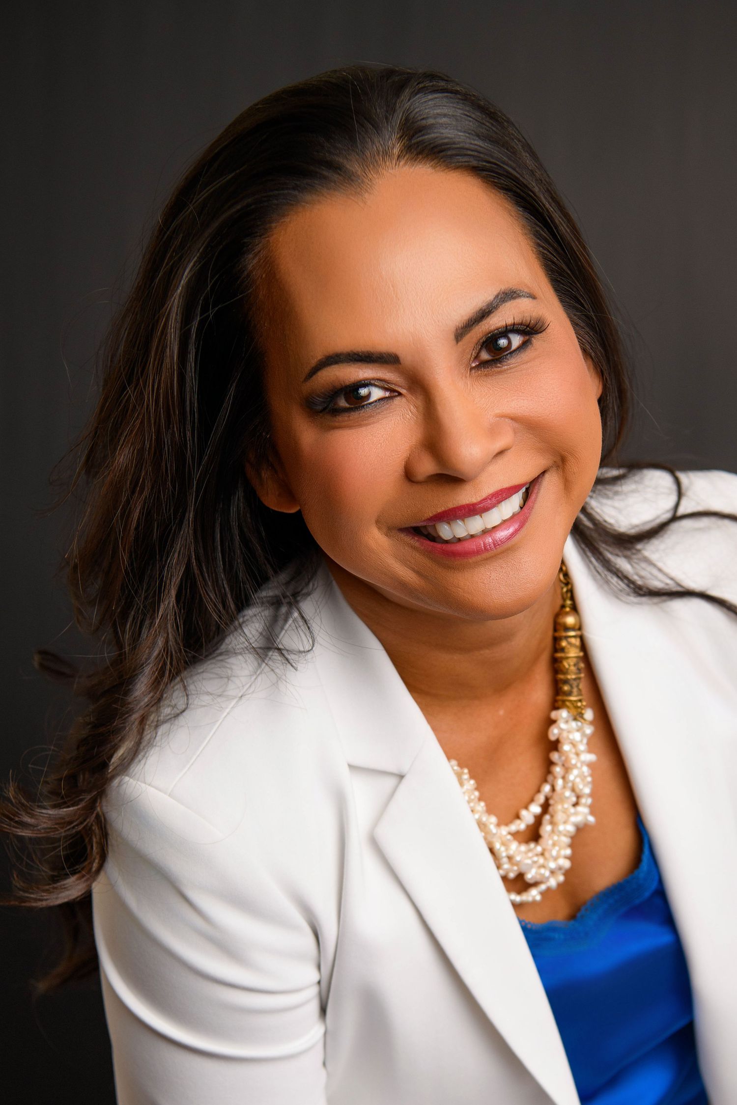 Professional headshot portrait of a person in a white blazer and blue top wearing a layered pearl necklace against dark background.