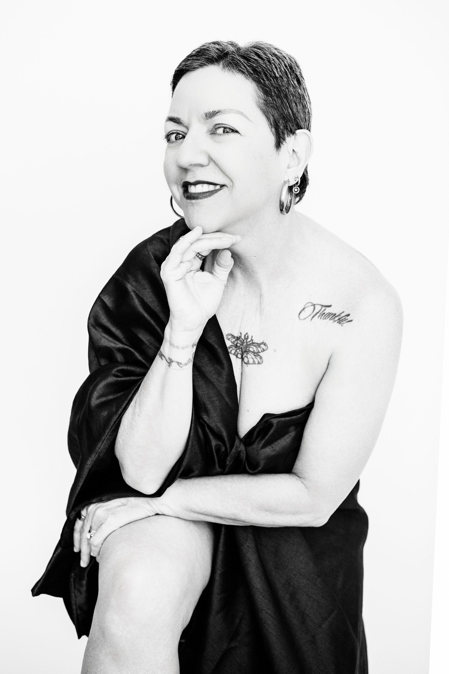 Black and white portrait of a person in black clothing with short hair and tattoos posing against white background.