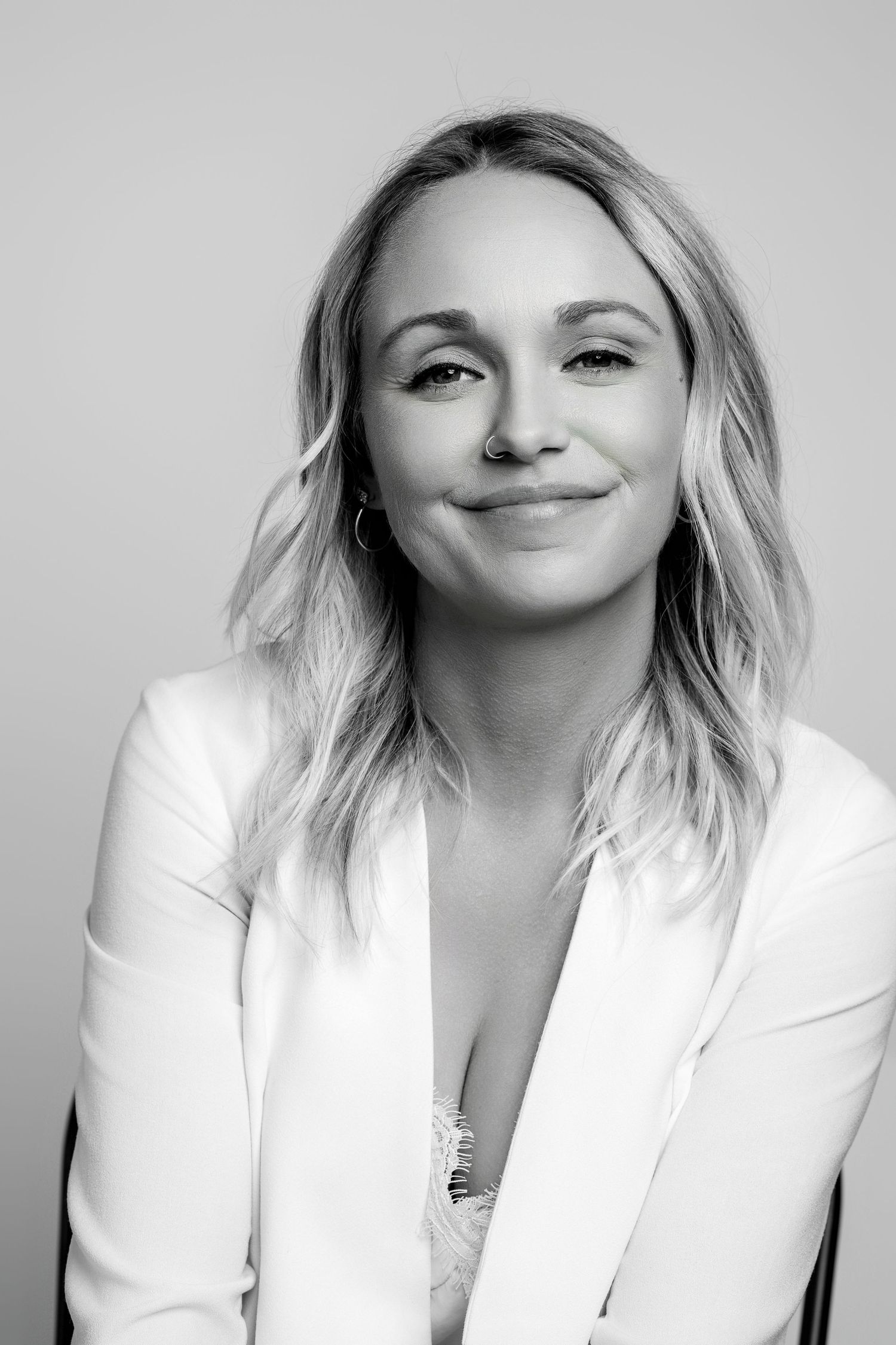 A black and white portrait of a smiling person wearing a white blazer with wavy blonde hair against a grey backdrop.