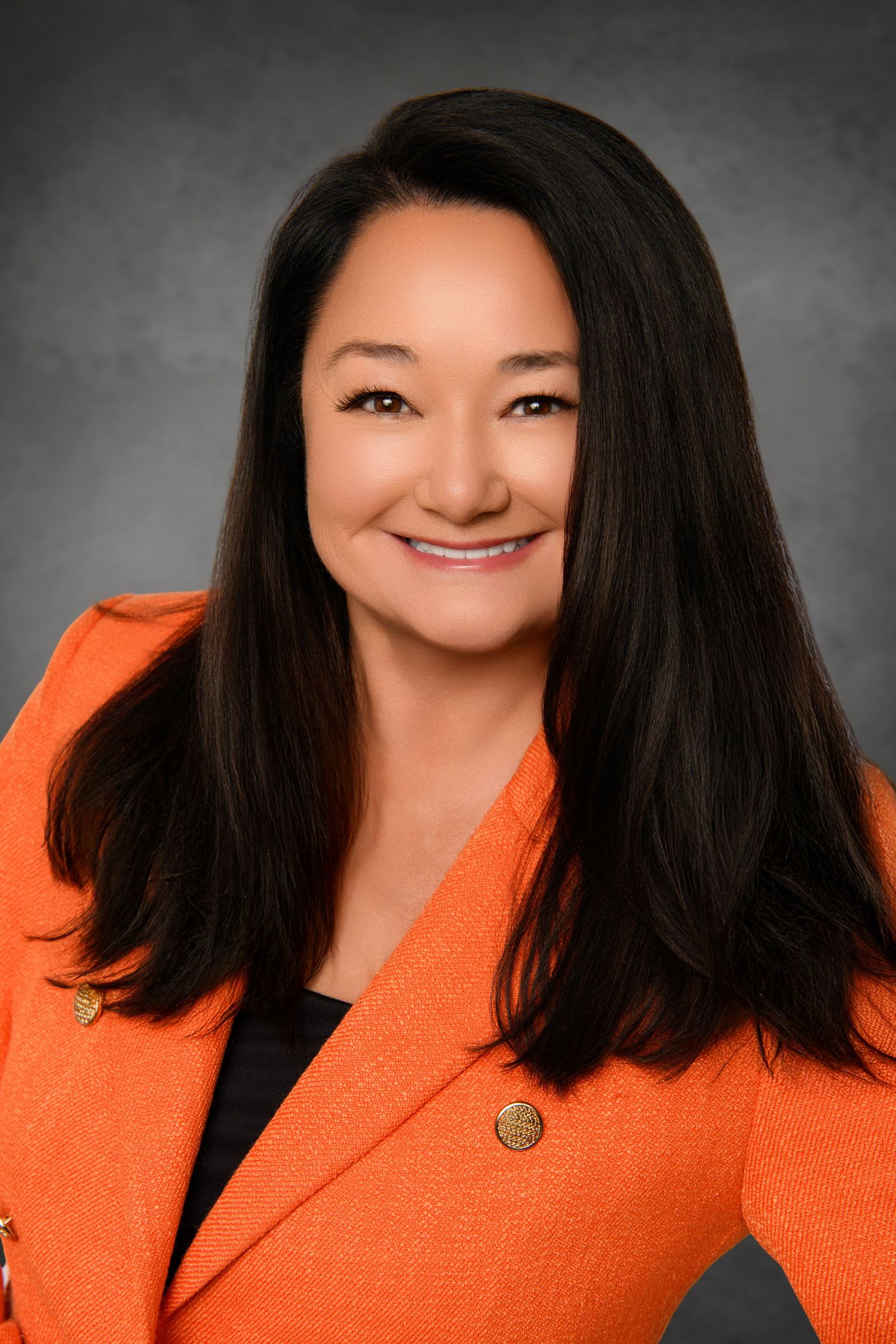 Professional headshot of a dark-haired person smiling warmly while wearing an orange blazer against a gray background.