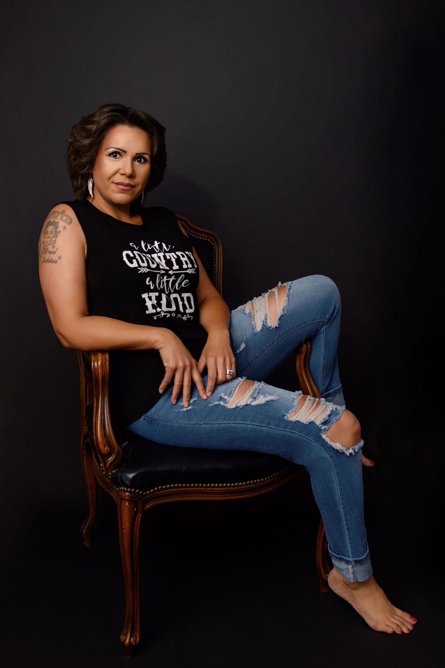 Woman in Pink Floyd t-shirt and ripped jeans poses casually on vintage chair against dark backdrop.