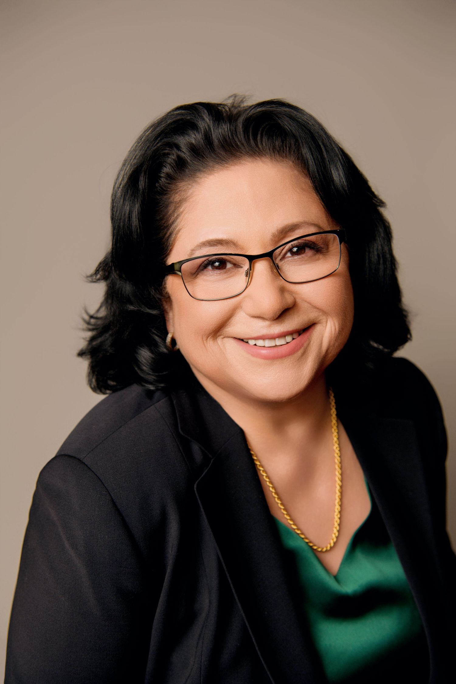Professional business headshot of a person with dark hair wearing glasses, a black blazer and emerald green top.