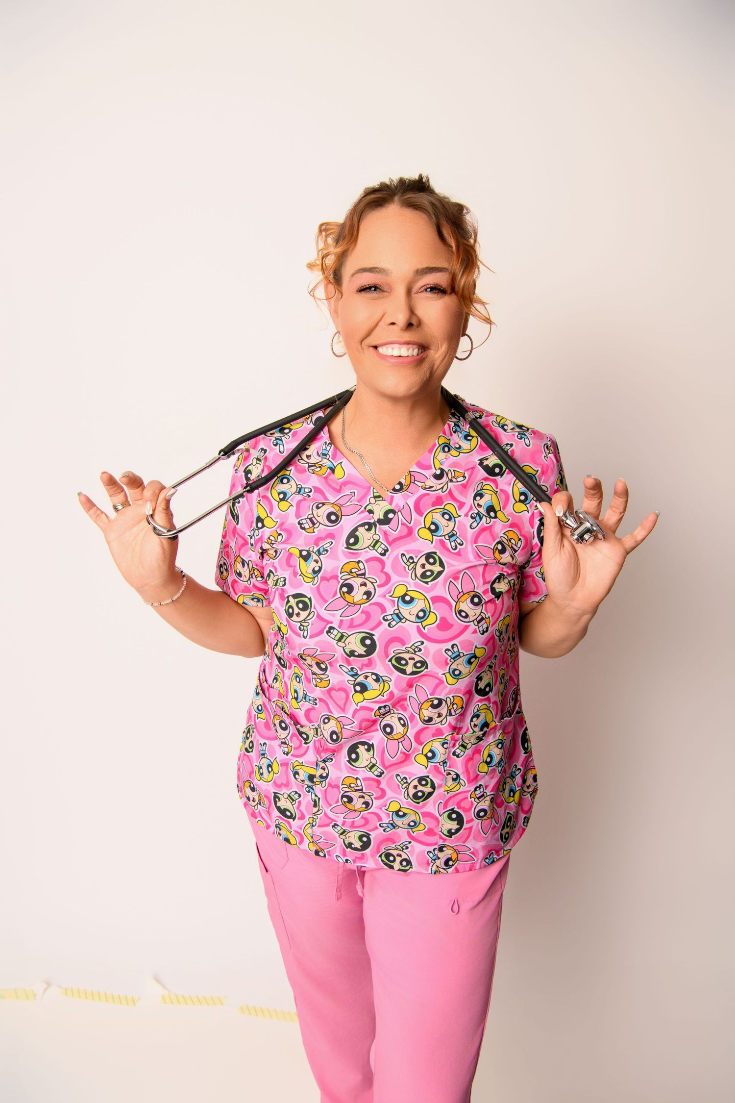 Healthcare worker wearing pink floral scrubs holds stethoscope while smiling against light background.
