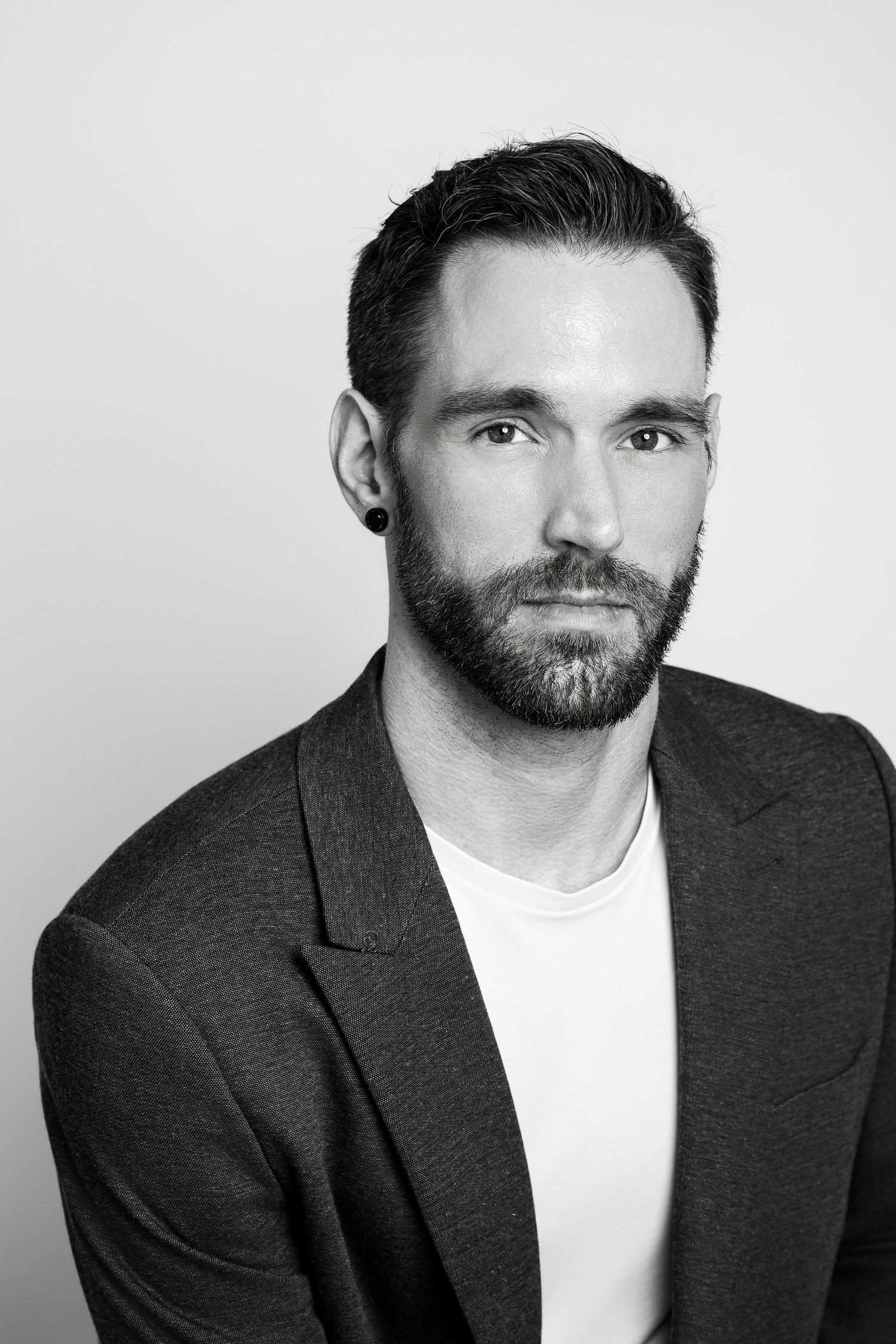 Black and white professional headshot of bearded man wearing dark blazer and white t-shirt against light background.