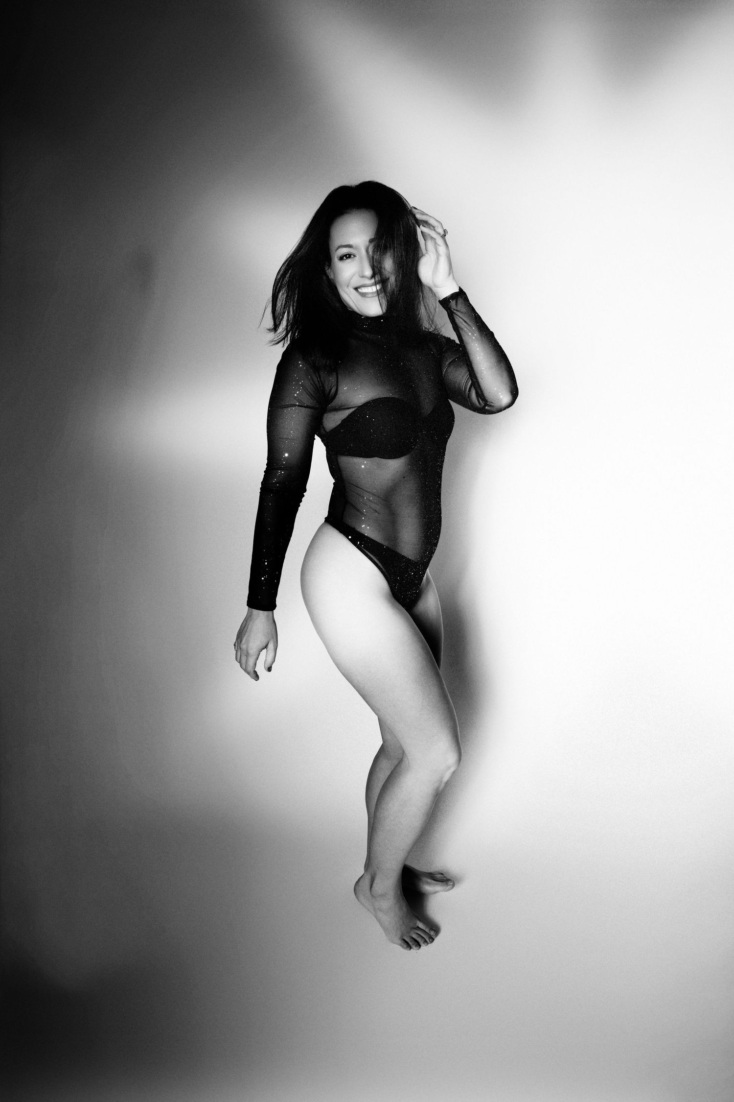 Dynamic black and white photo of model in mesh bodysuit captured in mid-motion against light background.