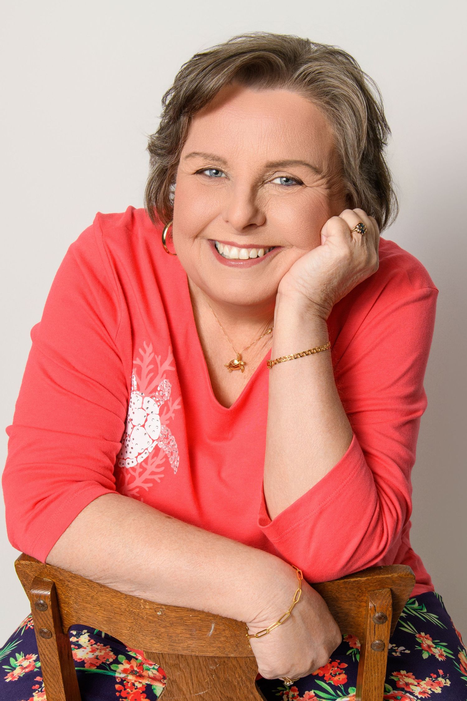 Casual portrait of person in coral top sitting on wooden chair with floral skirt.