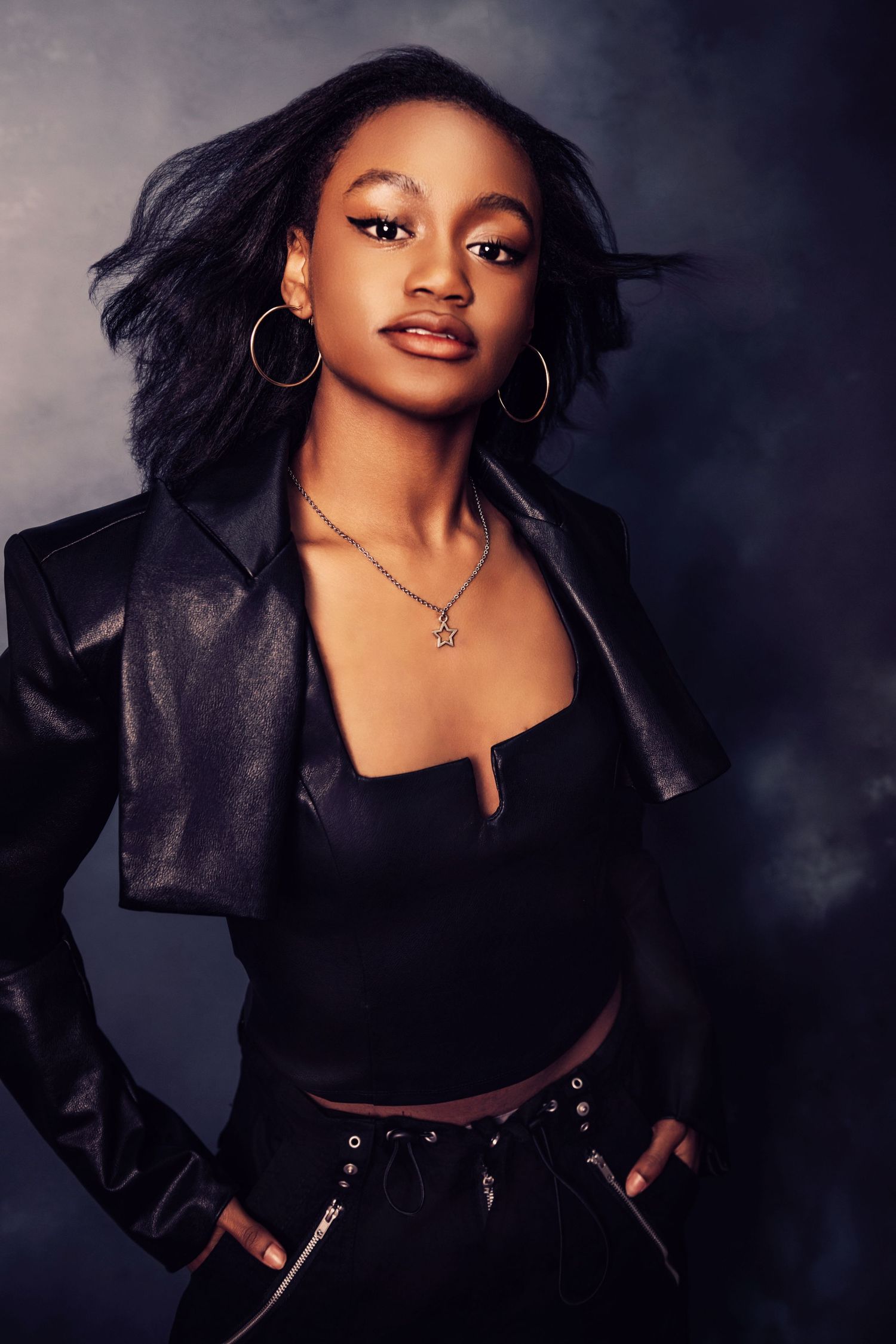 Studio portrait featuring black leather jacket and statement jewelry against dark backdrop.