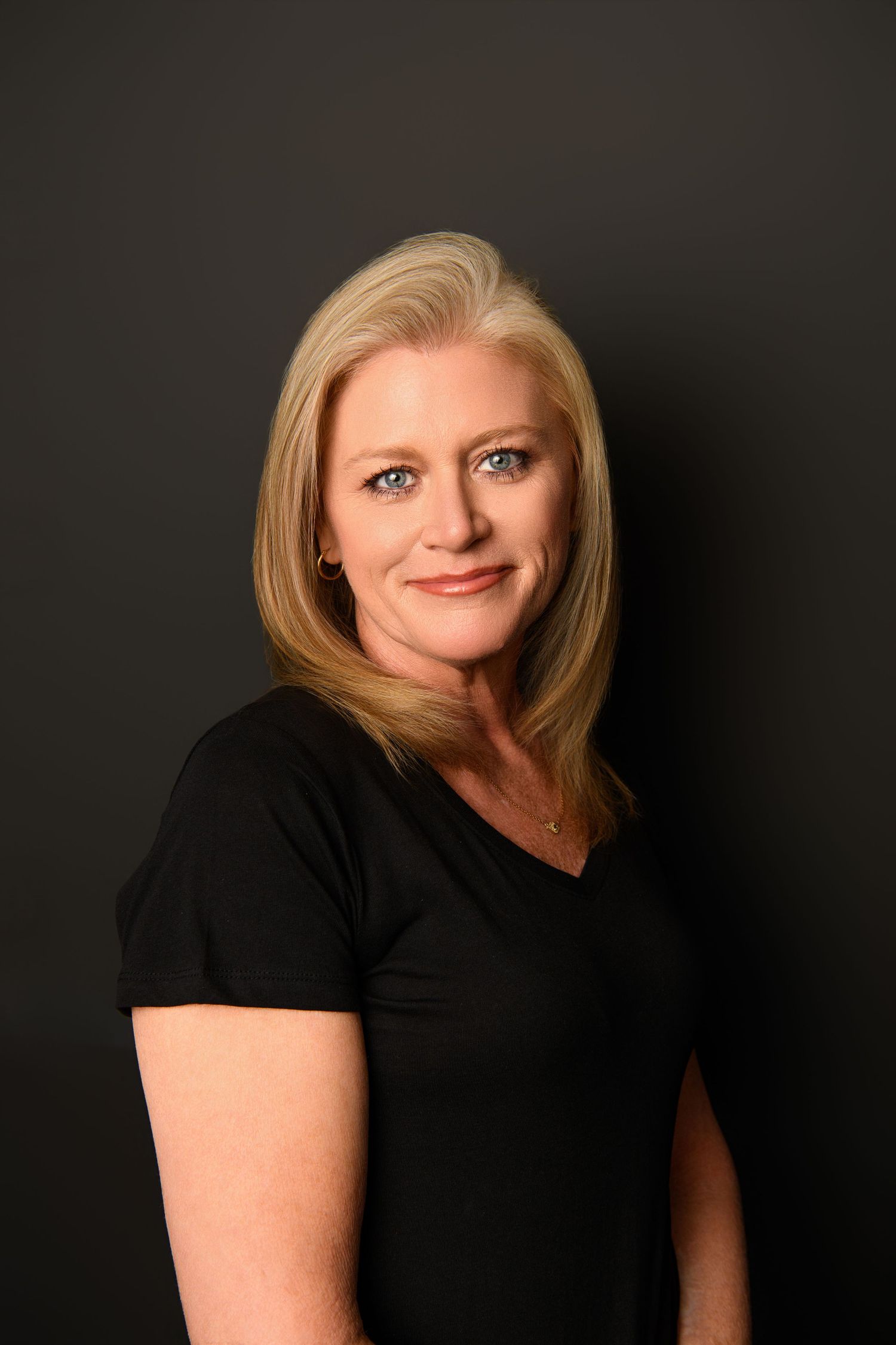 Studio portrait in black v-neck top with blonde hair against dark backdrop.
