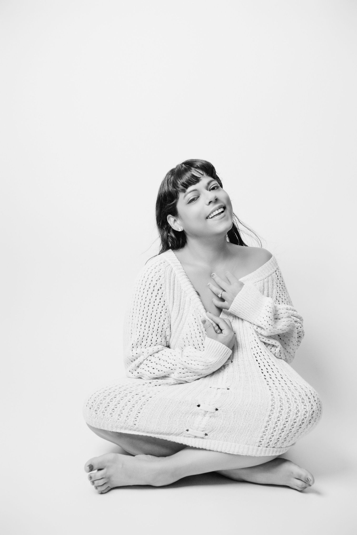 Casual portrait of a model in white knitwear sitting cross-legged on white background.