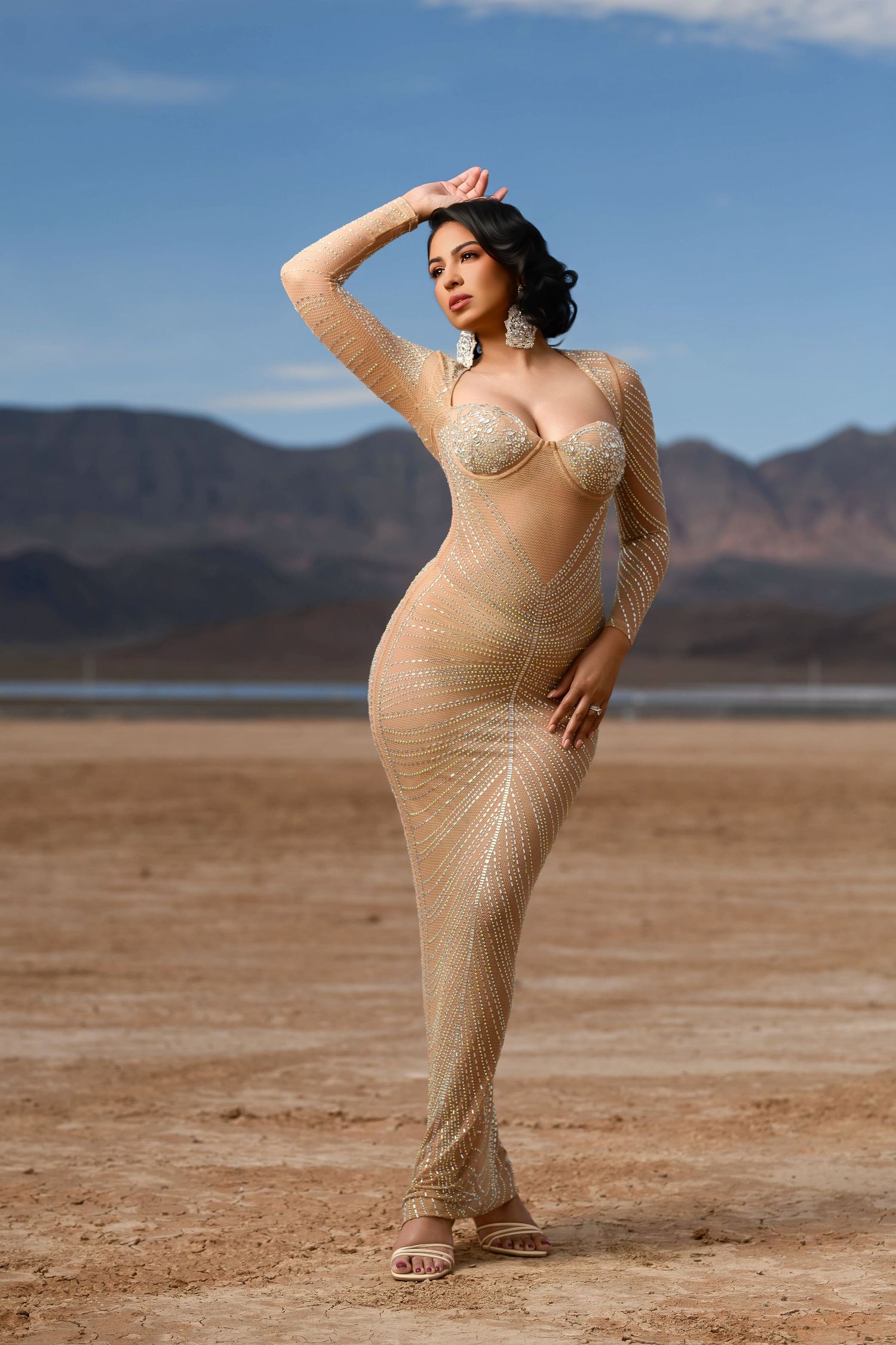 Desert photoshoot featuring figure in sheer beige dress with mountains in background.