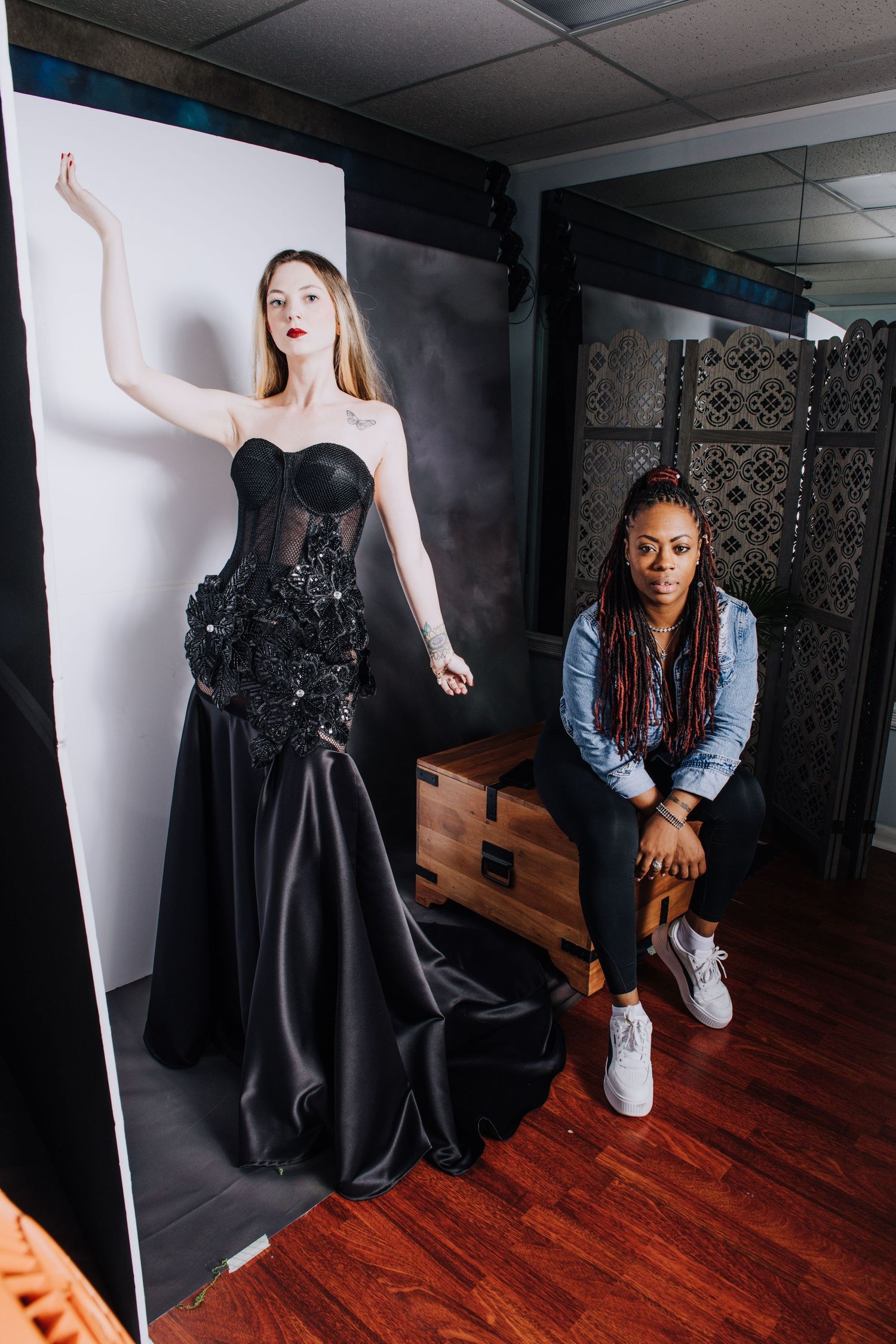 Behind-the-scenes photo showing a black evening gown fashion shoot setup with model and photographer in studio space.