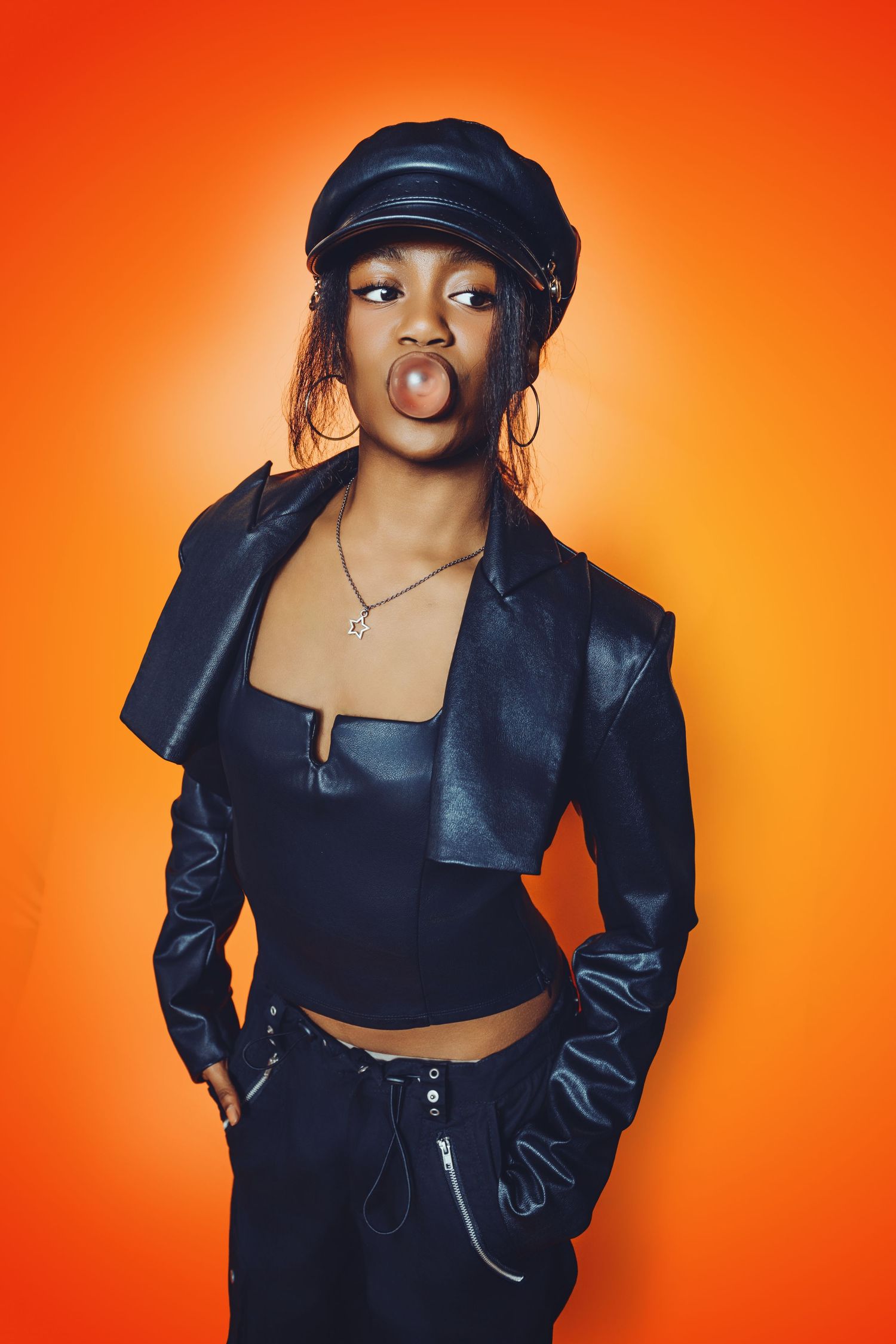 Playful portrait in leather jacket and cap against vibrant orange background.