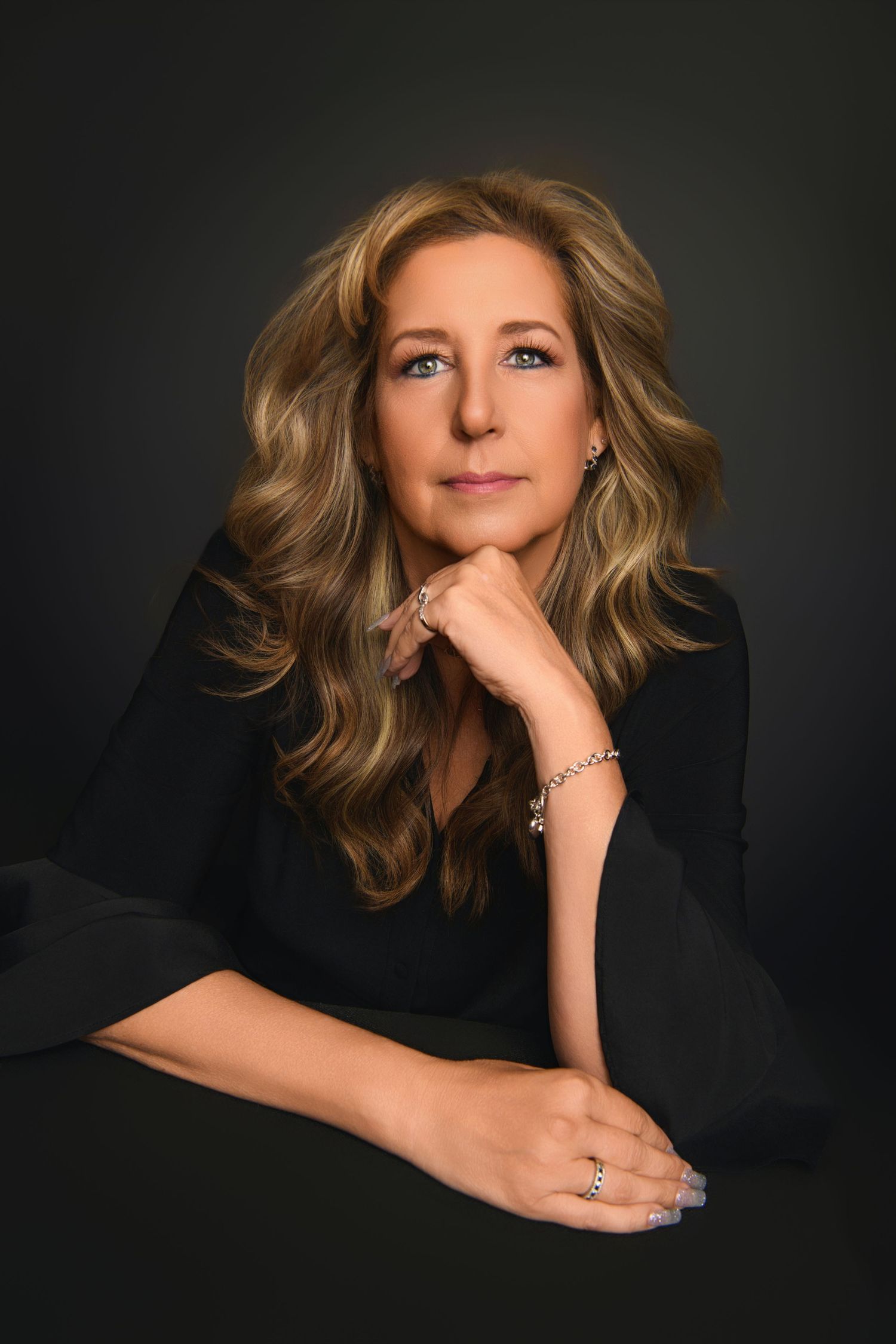 Professional portrait of woman in black blouse with wavy blonde hair against dark background.