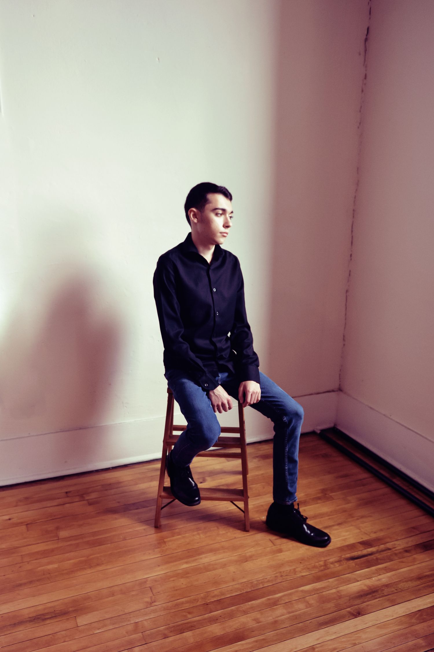 Professional studio portrait of person in dark button-up shirt and jeans sitting on wooden stool against white wall.