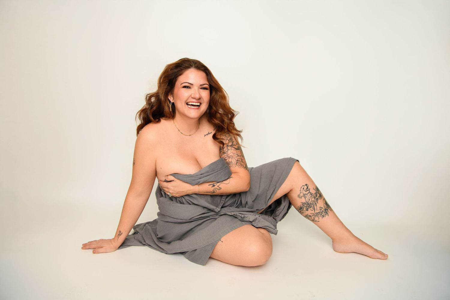 A person in a gray wrap dress poses cheerfully on a white backdrop with bare shoulders and visible tattoos.