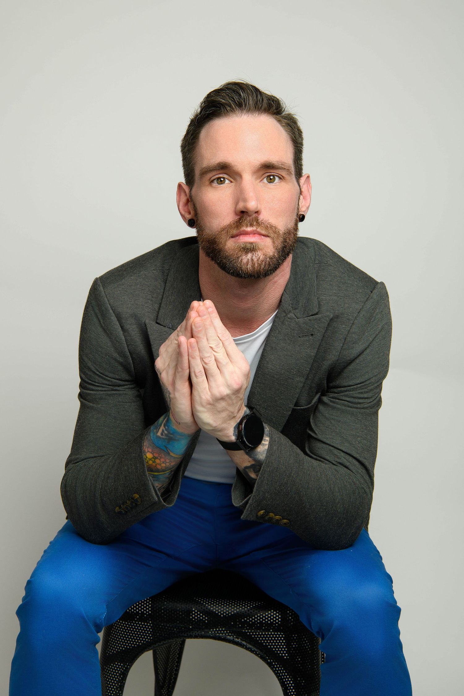 Portrait of person in gray blazer and blue pants sitting on stool with hands clasped in prayer position against white background.