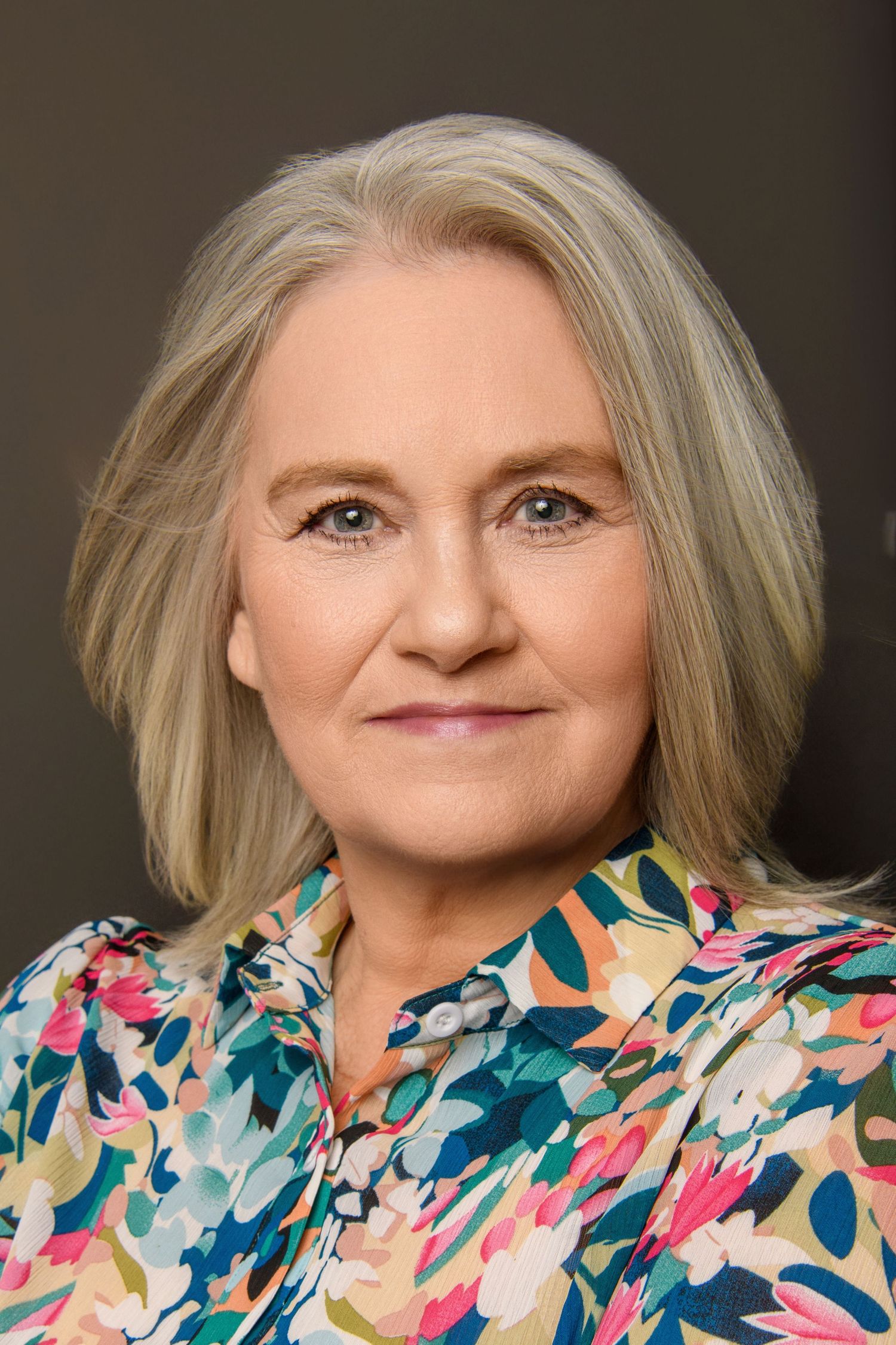 Professional headshot featuring short blonde hair and colorful floral print blouse.
