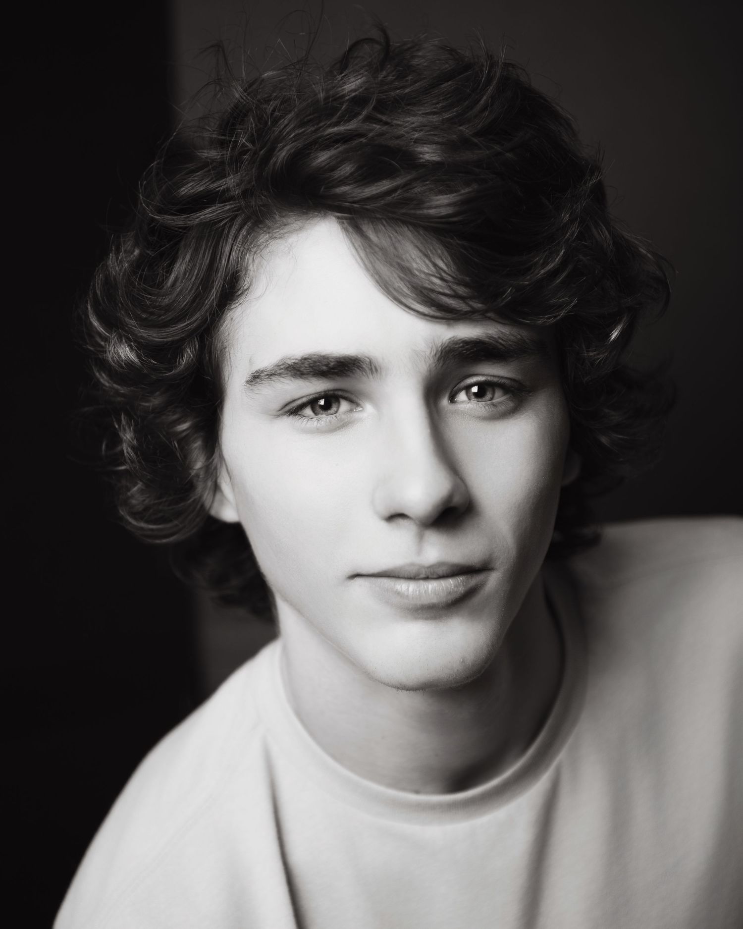 Portrait of a young man with dark curly hair wearing a white sweater against a dark background.