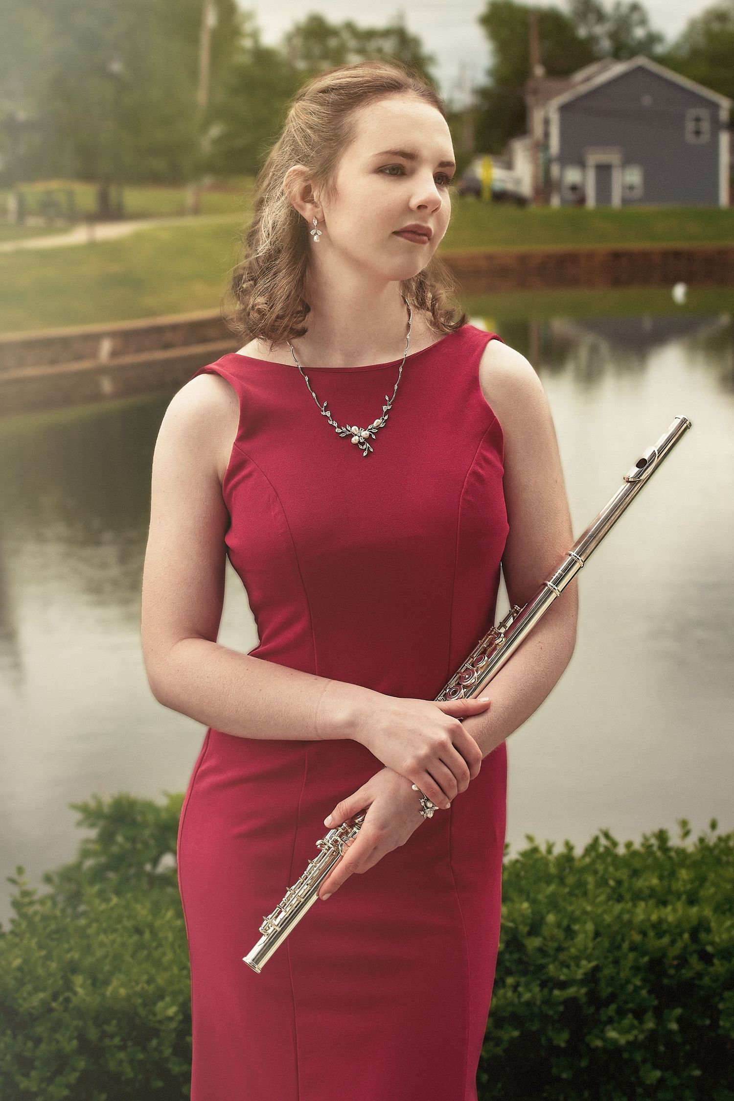 Musician in red formal dress poses outdoors with flute near water.