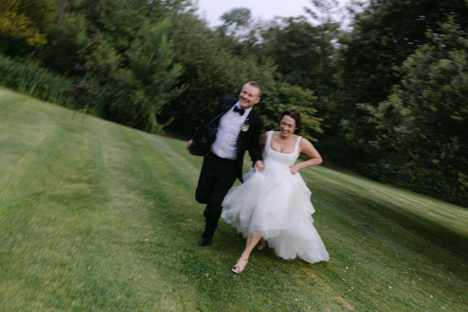 A couple runs playfully across a green hillside during their wedding photoshoot.